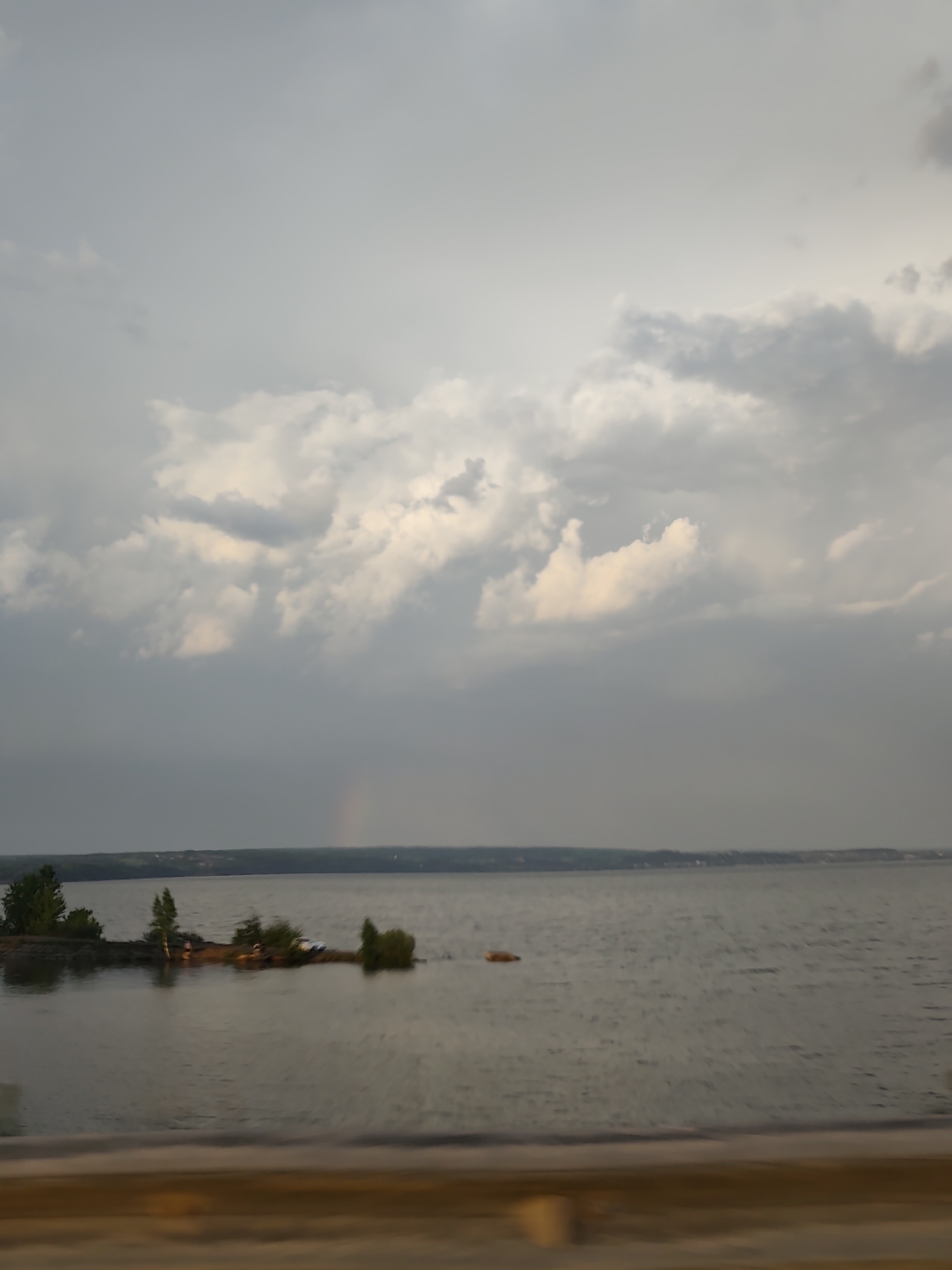 The image shows a body of water, possibly a lake or river, under an overcast sky with clouds. In the distance, there is a land mass with a visible horizon line. A small, tree-covered island is located in the water. The location is Пальники, Russia. A faint rainbow is discernible in the sky. The foreground is out of focus, suggesting the image was taken from a moving vehicle or behind an object. No people are clearly visible.
