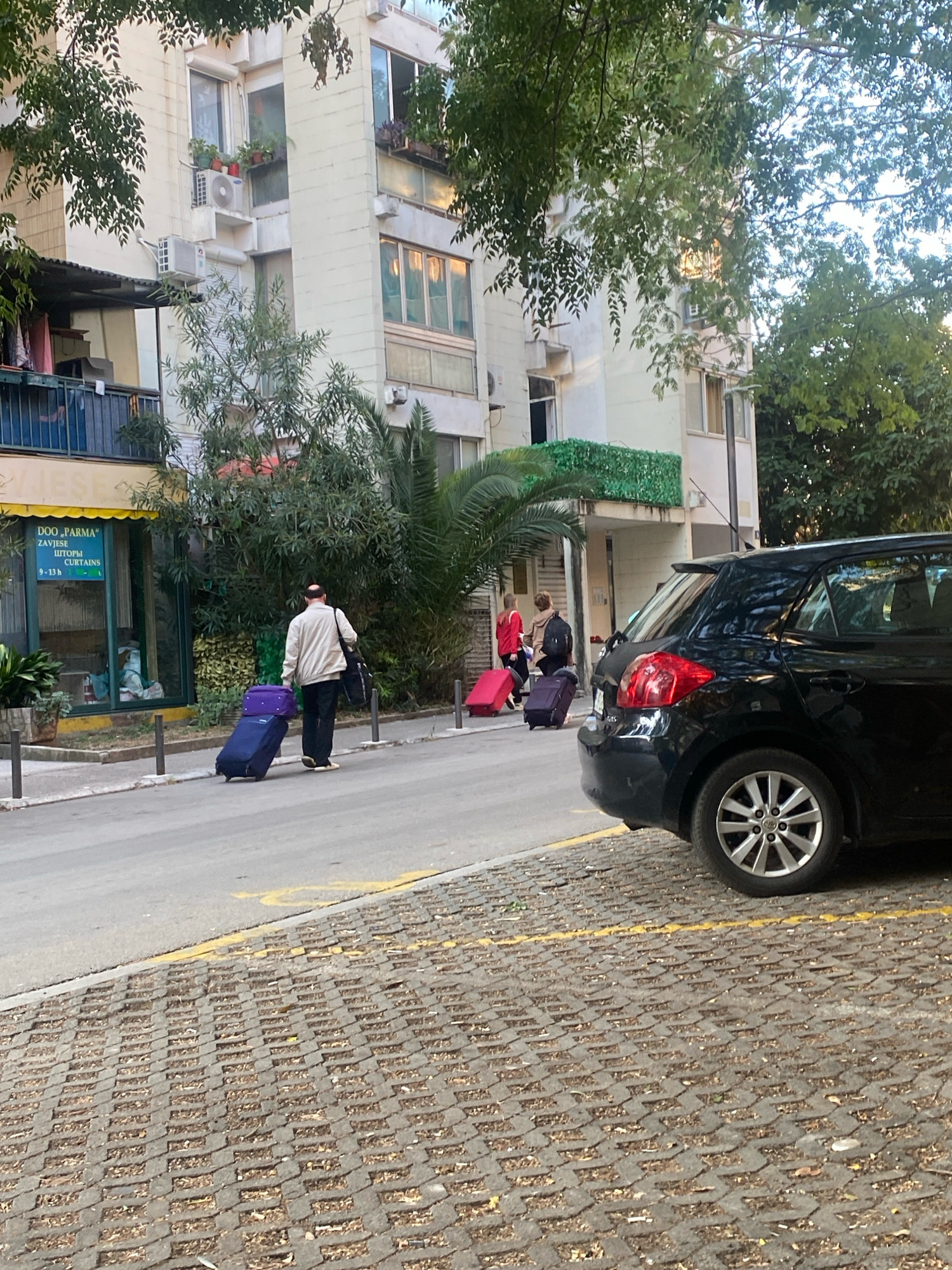 The image captures a street scene in Budva, Montenegro. In the foreground, a black hatchback car is parked on a cobblestone lot next to a paved street. The street has yellow lines indicating parking or traffic direction. On the street, a man is walking away from the camera, pulling a dark blue rolling suitcase with a purple bag on top. Further down the street, two women are walking towards the camera, one pulling a red rolling suitcase and the other pulling a dark blue rolling suitcase. Behind the street and sidewalk is a multi-story building with balconies and windows, flanked by trees. A storefront with a yellow awning is visible on the left side of the building, displaying signage. The overall impression is of a typical urban environment with people engaged in travel or daily activities.