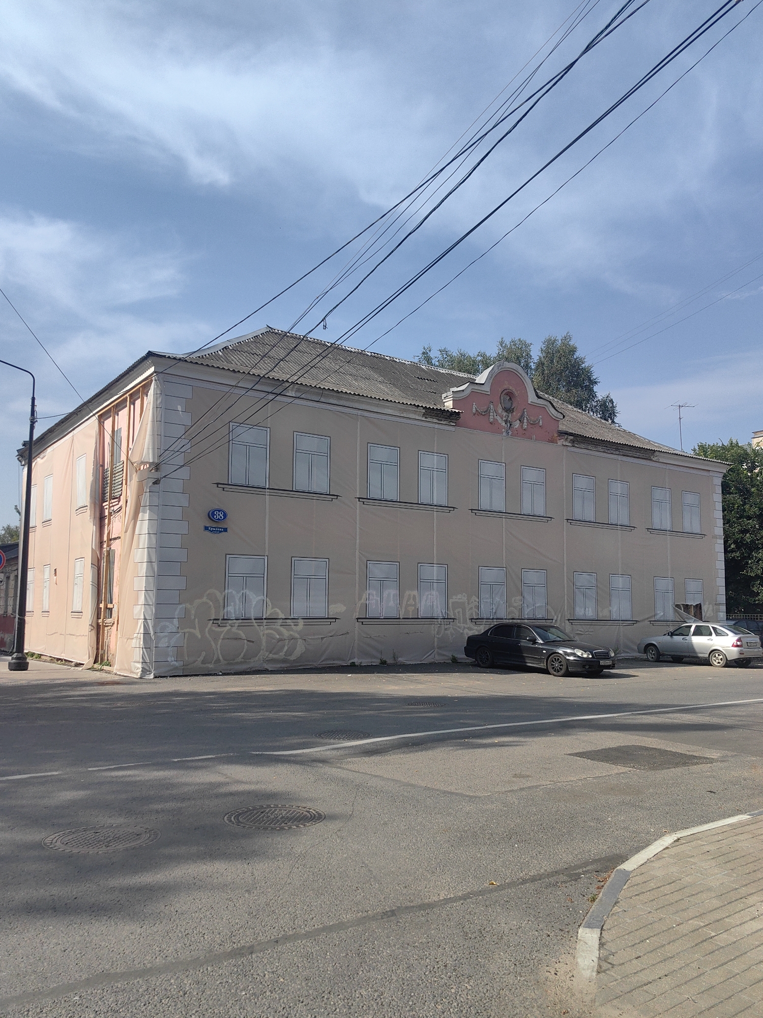 The image shows a two-story building covered in scaffolding and a light-colored tarp, with a grey corrugated roof. The building appears to be under renovation. The windows are outlined with white frames, and some graffiti is visible on the lower part of the tarp. A blue street sign with the number "38" and text that reads "Крылова" (Krylova) is mounted on the building's facade, indicating its location on Krylova Street in Tver, Russia.
In front of the building, a paved road is visible, with two cars parked on the side: a black sedan and a silver sedan. The road has lane markings and two manhole covers. The environment is sunny with a blue sky and scattered clouds. Trees are visible in the background. The overall scene depicts an urban street with a building undergoing renovation.