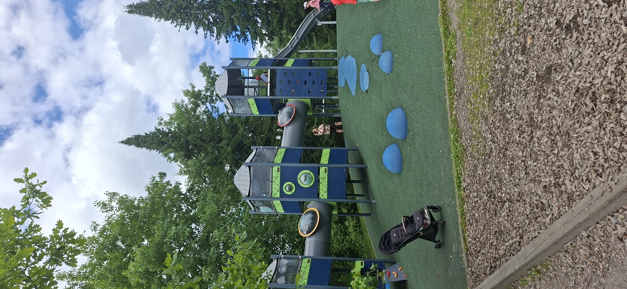 An outdoor playground with a large, multi-level play structure featuring slides, tunnels, and climbing elements is depicted. The play structure is primarily blue and green with gray accents. The ground surface is a bright green, artificial turf with several blue, dome-shaped elements scattered on it, resembling large pebbles or stepping stones. A black stroller is parked on the left side of the play area. In the background, a dense line of green trees forms a natural backdrop, and a cloudy sky with patches of blue is visible above. There are at least three people visible: a woman standing with her arms crossed on the play structure, a child in a red hat on a higher level of the play structure, and a child in a pink hat near the slide. The playground is situated in an area with a border of wood chips and gravel. Based on the image content and surrounding context, this playground appears to be located in Lahti, Finland.