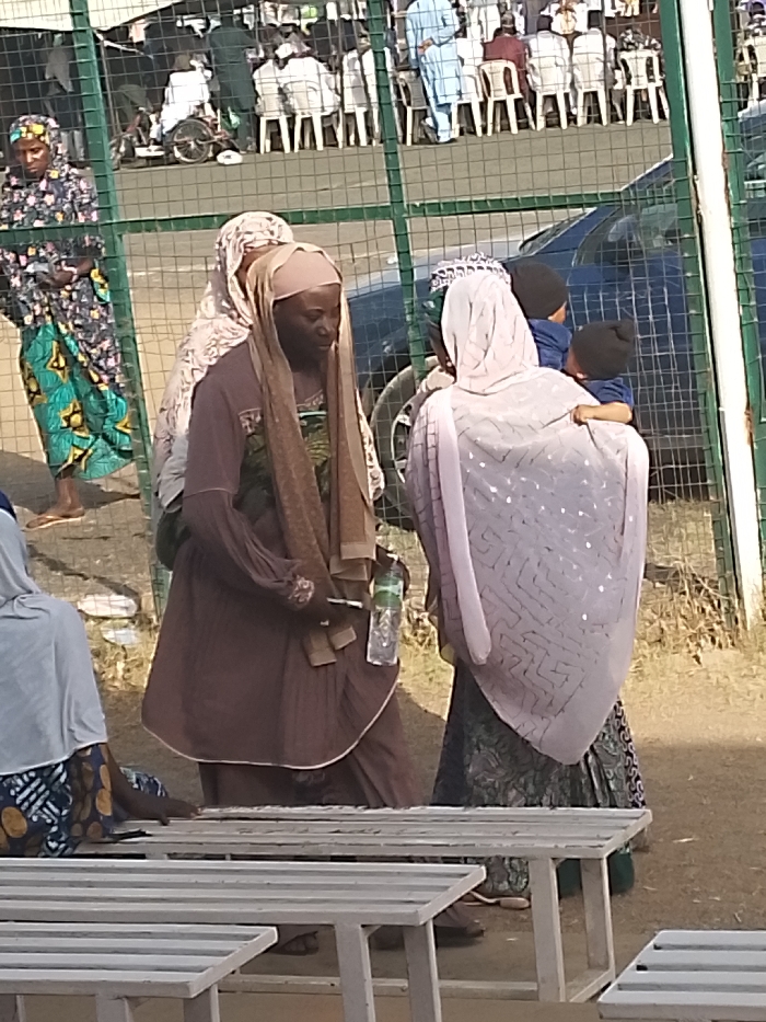 This image depicts an outdoor gathering in Sabon Gari East, Nigeria. In the foreground, a light-colored slatted bench is partially visible, with a person seated next to it, wearing a light headscarf and patterned skirt.
The central focus includes three women standing in the midground. One woman, with a darker complexion, wears a light brown hijab and a long-sleeved brown tunic, holding a clear plastic water bottle in her right hand and a small, dark object in her left. She is partially obscured by another woman wearing a patterned headscarf directly behind her. To their right, a third woman faces away from the viewer, clad in a light, patterned head covering and a dark, patterned skirt. She holds two small children, both wearing dark beanies and blue clothing.
A green wire mesh fence separates the midground subjects from the background. Behind this fence, a dark-colored sedan is parked. Further in the background, numerous individuals, primarily men in light-colored traditional attire and head coverings, are seated on white plastic chairs. A person in light blue stands among them, and a bicycle is visible near the seated group. The ground appears dry and dusty under daylight conditions.