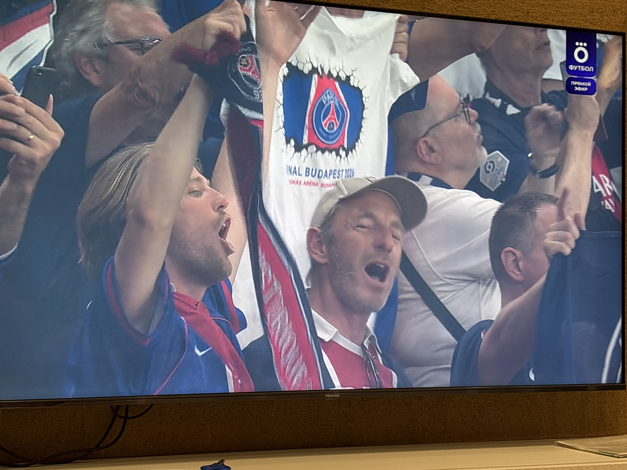 The image appears to be a photograph of a television screen showing a sporting event. The scene displays several people, seemingly fans, cheering and engaged in supporting their team. Some are holding up scarves and wearing team apparel. A t-shirt is visible with the Paris Saint-Germain logo and text reading "Final Budapest 2026." The environment seems to be a stadium or similar large gathering space. The upper right corner of the television screen shows a logo and text written in Russian, including "ФУТБОЛ ПРЯМОЙ ЭФИР". The context suggests the event is likely a football (soccer) match. There is no indication that the image was taken in Votkinsk, Russia. The presence of Russian text on the TV screen, however, may indicate that the image was taken in Russia or a Russian-speaking region.