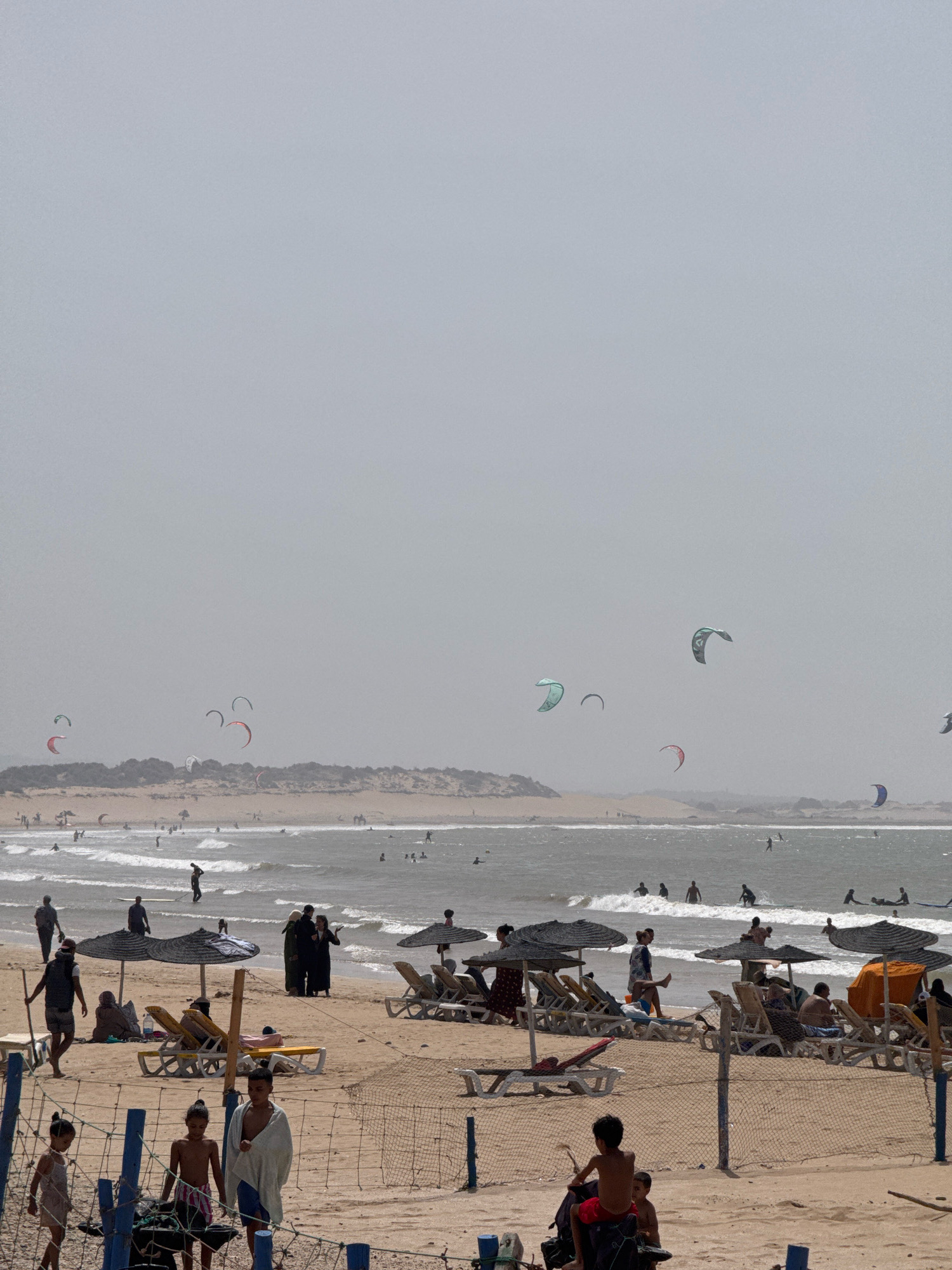 The image depicts a crowded beach scene in Essaouira, Morocco, characterized by a hazy, overcast sky. The foreground is dominated by a sandy beach with several people, including children and adults, visible. Some individuals are walking along the shore, while others are relaxing on lounge chairs under umbrellas. A net fence runs across the lower portion of the image, separating the beachgoers from the foreground. In the middle ground, a line of umbrellas and lounge chairs provides shade. The ocean is visible with moderate waves, and several individuals are in the water, swimming or participating in water sports. Notably, there are numerous kitesurfers in the ocean, their colorful kites dotting the sky and water. The background features a strip of sand dunes covered with sparse vegetation, characteristic of the coastal landscape of Essaouira. The overall impression is a busy day at a popular beach destination.