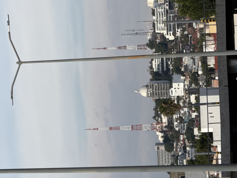 The image features an urban landscape with multiple buildings and towers. A tall, red-and-white striped communications tower is prominent. Other towers of varying heights are visible in the distance. Residential and commercial buildings are densely packed together. A curved, gray lamppost is in the foreground on the left. The sky is overcast.