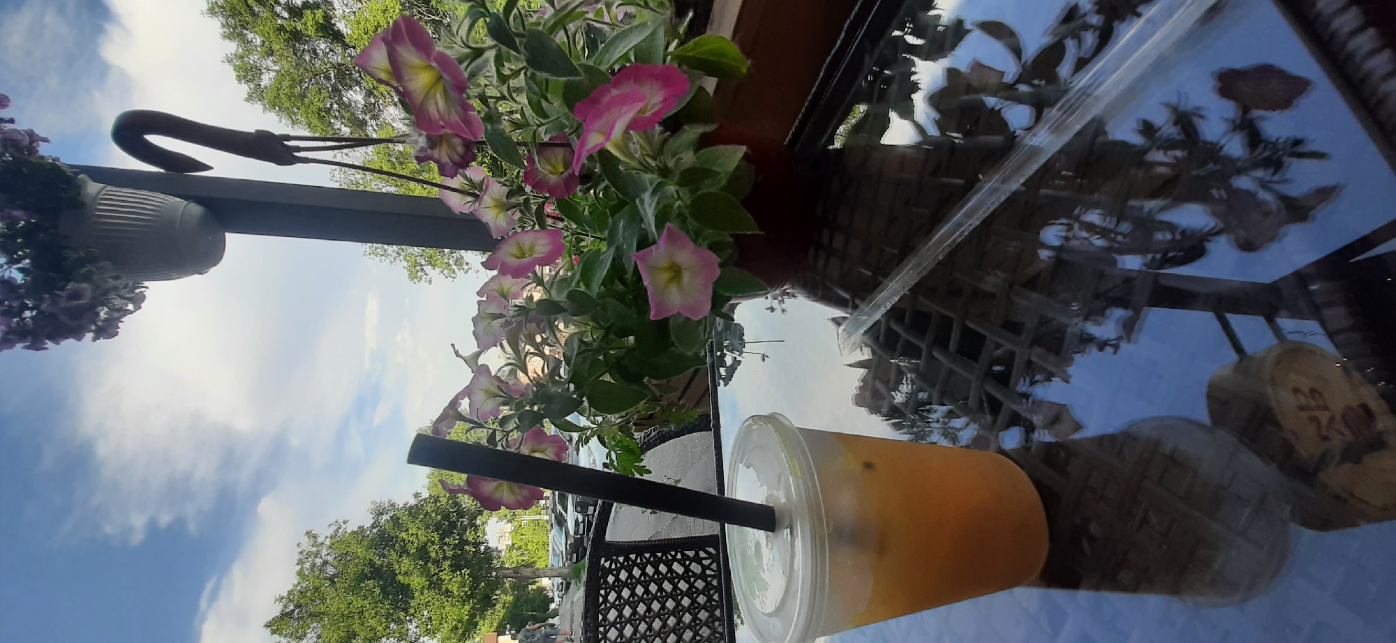 A clear plastic cup filled with an orange liquid and a black straw sits on a reflective glass table. In the background, pink and yellow petunias bloom in a planter, with a blurred street scene and trees visible beyond. The location is Yessentuki, Russia, a resort city known for its mineral springs and spa culture. The scene suggests a cafe or outdoor seating area, possibly a place for tourists to relax and enjoy a beverage while taking in the surroundings. The reflection on the table shows the intricate pattern of the table itself, as well as the flora and sky above, adding depth and visual interest to the composition.