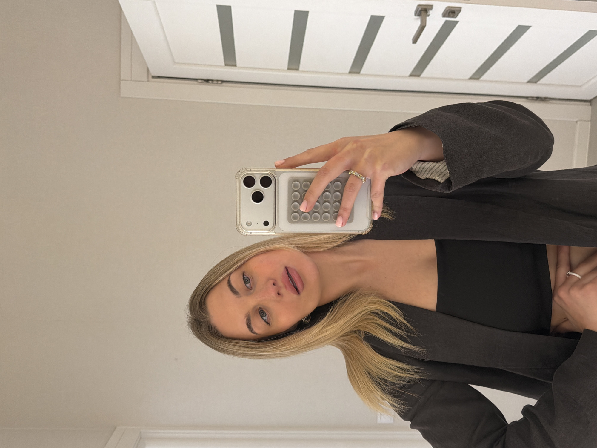 This is a mirror selfie taken indoors, featuring a young woman holding a smartphone. She has long, straight blonde hair, fair skin, and light-colored eyes. Her expression is neutral to slightly pensive, with her lips slightly parted. She is wearing a black crop top or bra under a dark, possibly charcoal-colored, jacket or blazer, which has a striped lining visible at the cuff of her right sleeve.
She holds a clear-cased iPhone, likely a Pro model due to the camera array, with a white or light gray pop-it style accessory on its back. On her right hand, visible in the selfie, she wears at least two rings: one on her ring finger and another on her middle finger, both appearing to be jeweled. Another ring is visible on her left hand's ring finger. Her fingernails have light pink polish.
The background consists of a light-colored wall and a white door with decorative vertical panels and a metal handle. The lighting appears bright and even, suggesting an indoor setting during the daytime. There is no visible text in the image. The location context provided is Sofiivska Borshchahivka, Ukraine.