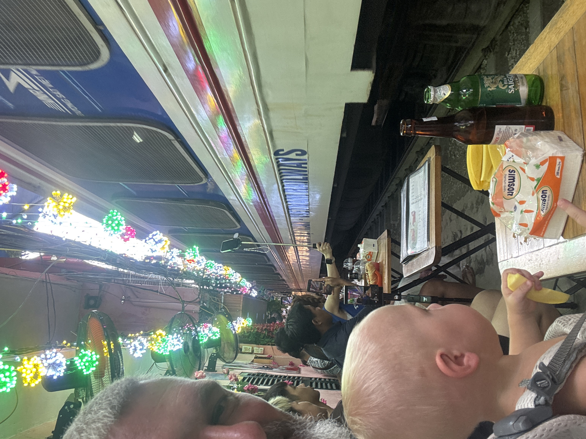 This image captures a bustling scene at a cafe on Train Street in Hà Nội, Vietnam, as a train passes by. In the immediate foreground on the left, the profile of an adult male with a gray beard is partially visible, looking towards the right. Below him and slightly to the right, a blonde-haired toddler or baby sits in a carrier or high chair, holding a yellow object, possibly a piece of fruit or toy, and gazing towards the right.
On a wooden table in the lower right, two beer bottles are present: one brown bottle and one green bottle labeled "BIA SAIGON SPECIAL." Next to them, a package of "SIMSON" brand napkins with a carrot design is visible. Further back, on a different table, some yellow items resembling fruit or snack packaging are present.
The midground features several individuals seated at cafe tables arranged along active train tracks. A woman on the far right holds a selfie stick with a smartphone, extending it towards the passing train to capture a photo or video. Another person to her left is also holding a smartphone, seemingly recording. Other patrons are seated at adjacent tables, observing the train.
Dominating the right side of the frame is a blue and white train car, positioned extremely close to the cafe tables. The words "VIETNAM RAILWAYS" are clearly visible in blue lettering on the white section of the train car. Above this, near the top of the train, text in Vietnamese reads "ĐƯỜNG SẮT VIỆT NAM" (Vietnam Railways) with an accompanying logo. The train tracks are visible directly beneath the train and extending under the cafe tables.
Above the scene, numerous strings of decorative, multicolored LED lights, shaped like stars or flowers (green, yellow, white, red, pink), illuminate the narrow passage. Several ceiling fans are also mounted overhead amongst the lighting and extensive electrical wiring. The background on the left shows the facade of a building, possibly part of the cafe, decorated with artificial flowers and vines.