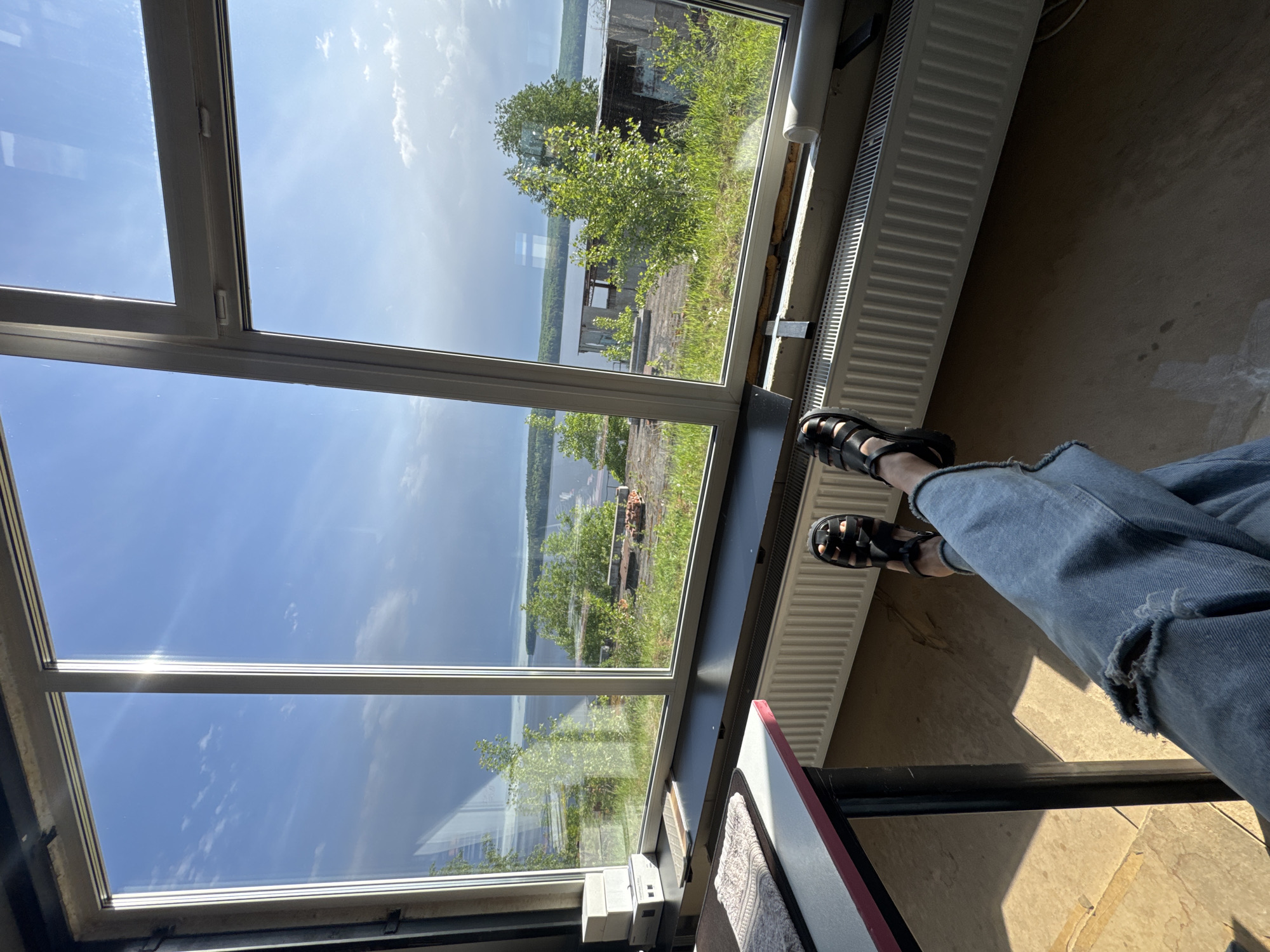 The image shows a person seated by a large window, likely indoors. The window offers a view of a body of water, possibly the Kama River, and the opposite shore. Trees and some buildings are visible on the far bank. The person is wearing blue jeans with a ripped knee and black sandals. A radiator runs along the base of the window. A small table or shelf with items on it is located next to the person. The scene's location is Chaikovsky, Russia, based on the landscape.