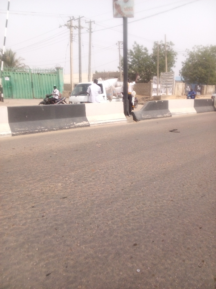 The image depicts a roadside scene in Sabon Gari East, Nigeria, under bright, possibly hazy daylight conditions. The foreground consists of a worn asphalt road surface. Two concrete barriers, painted white with black tops, run horizontally across the midground, separating the road from an adjacent area. A dark sandal or flip-flop lies on the asphalt to the right of these barriers.
Beyond the barriers, from left to right: a large green metal gate, partially open, is visible with a striped barrier arm extending from it. A dark motorcycle is positioned in front of the gate, with a male individual wearing a light garment and a cap seated on it. To the right of the motorcycle is a white utility vehicle, possibly a minivan or small truck, with a license plate "AA 487 YY." The vehicle's rear cargo area is loaded with multiple sacks. A male individual in a white garment stands by the passenger side of this white vehicle.
Further to the right, a tall black pole with an advertisement or sign mounted at its top extends upwards. Another male individual, wearing a yellow shirt and dark trousers, stands behind this pole with his right arm raised. In the background, multiple wooden utility poles with numerous overhead power lines are visible. Sparse trees and light-colored buildings are present throughout the background. A male individual in a blue garment is seated on a low wall or structure on the far right, where a sign partially reads "NGK DAVID."