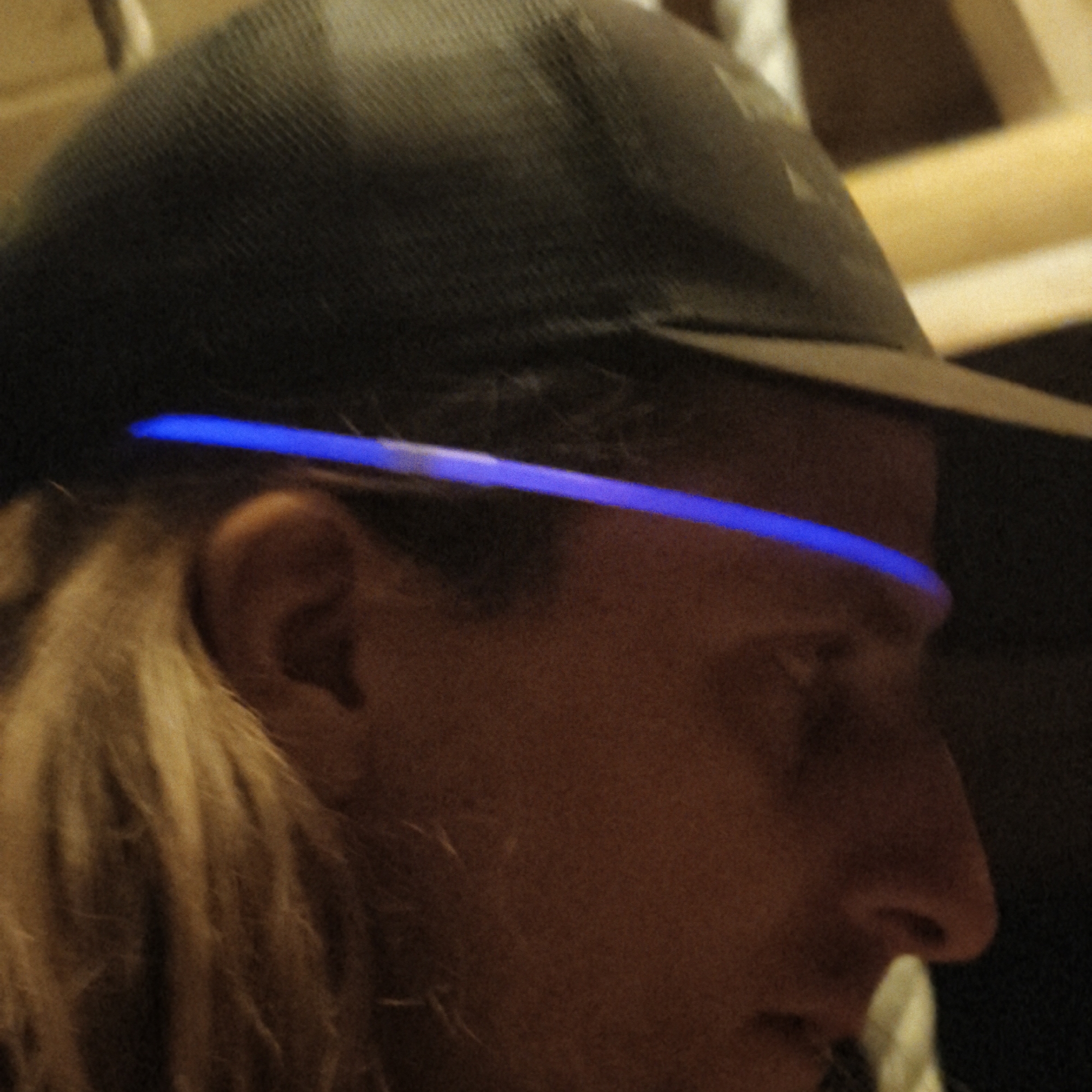 Close-up view of a person wearing a dark baseball cap and a blue light strip across their forehead. The person's face is partially visible, with a focus on the eye, nose, and ear. Blond hair is visible in the background. The image appears to be taken indoors, and there is no specific information to place this image within Капцёўскі сельскі Савет, Belarus, or any other location.