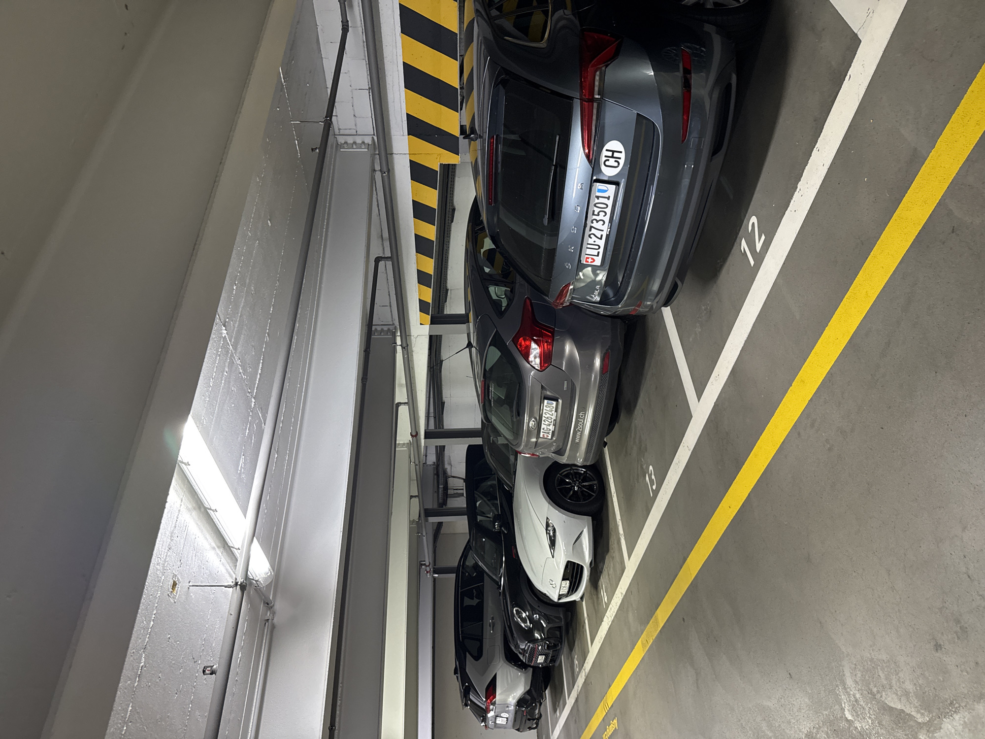 This media file shows a row of cars parked in an indoor parking garage. The cars are parked in designated spots marked with white and yellow lines. The scene is well-lit, suggesting it is daytime, although it is indoors. The setting appears to be a modern parking facility with a concrete ceiling and metallic structures.
The subjects are primarily the cars, which are of various makes and models, and appear to be in good condition. There are no people visible in the frame.
Notable details include the license plates of some of the cars. One license plate reads "LU-273501" with a "CH" sticker, indicating it is registered in Switzerland. Another license plate is visible as "AG 4262488". The parking spots are numbered, with "12" and "13" clearly visible. There are also hazard stripes, typical of parking garages, on the wall.
The image has been rotated, making it appear as if the cars are parked sideways. The location is indicated to be Lucerne, Switzerland, which is consistent with the visible license plates. There are no other overt location cues, such as signage or landmarks. The overall impression is one of order and organization within the parking structure.