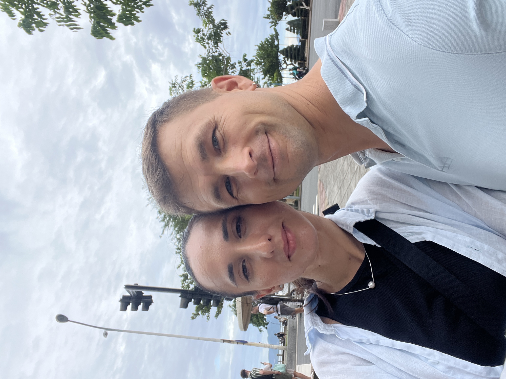 The image shows a selfie of a man and a woman in Nha Trang Ward, Vietnam. The man is fair-skinned with short brown hair. He wears a light blue polo shirt. The woman is fair-skinned with dark hair pulled back. She wears a black top, a light blue button-down shirt, and a pearl necklace. They are close together, with the man's head slightly above the woman's. Behind them, there is a street with trees, a traffic light, and pedestrians. The sky is cloudy. In the background, a waterfront is visible.