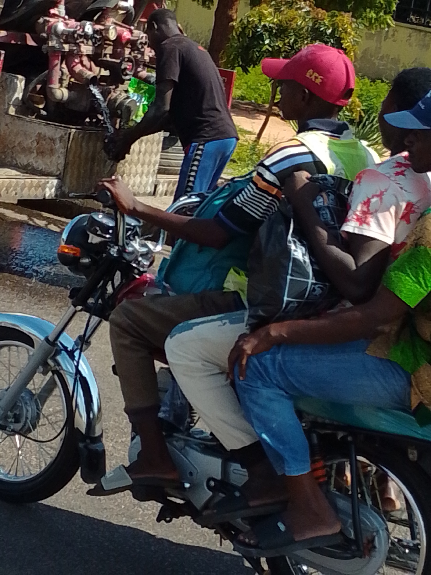 In Gombe, Nigeria, a man wearing a black t-shirt and blue shorts is operating a motorcycle. He is seen from the chest down, with his hands on the handlebars and his feet on the footpegs. He is looking forward, presumably driving. On the back of the motorcycle, there are three passengers. The passenger behind him is wearing a red baseball cap and a striped t-shirt with a yellow vest. The passenger behind that individual is wearing a white t-shirt with red floral patterns and a blue hat. The passenger furthest back is wearing a blue t-shirt and blue jeans. All passengers appear to be looking forward. The motorcycle is a red and black model with chrome accents. It is equipped with a rear rack and a chain guard. The scene appears to be a street in Gombe, with a red fire hydrant visible in the background. The road surface is asphalt. The overall impression is one of everyday life in a busy Nigerian city.