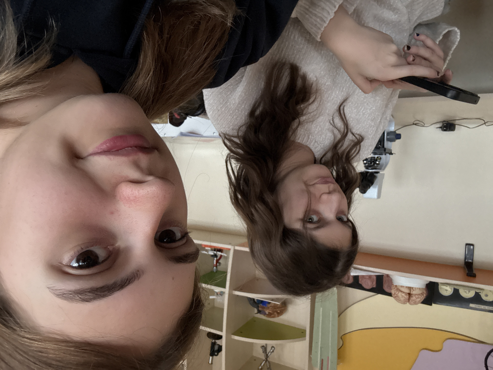 Two young women are posing for a selfie indoors. The woman on the left has light brown hair and is wearing a dark-colored top. The woman on the right has brown wavy hair and is wearing a light-colored sweater, she is holding a black smartphone. The background shows a classroom or science lab setting with shelves containing models and tools. A model of a human brain is also visible. While it is clear it's a classroom or lab, the exact location in Panagyurishte, Bulgaria, cannot be determined from the image alone.