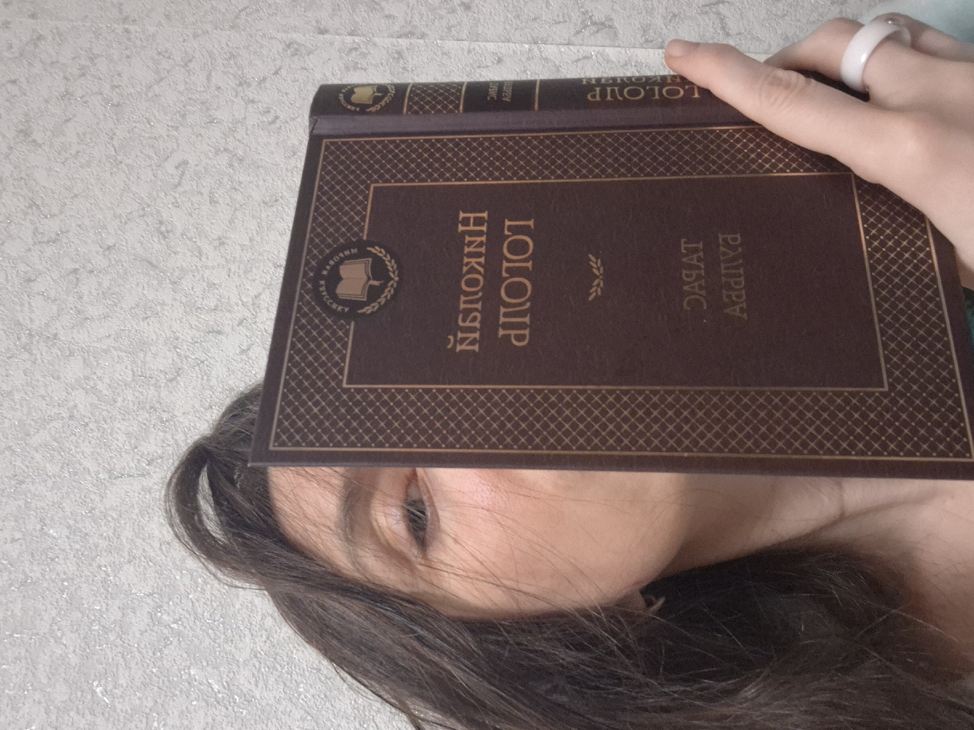 A person holds a book titled "LOLOLP Nikolaev" in Russian in front of their face. The person has dark brown hair. They are wearing a white ring on their right ring finger. The background is a textured white wall. The location is Unknown, Unknown.