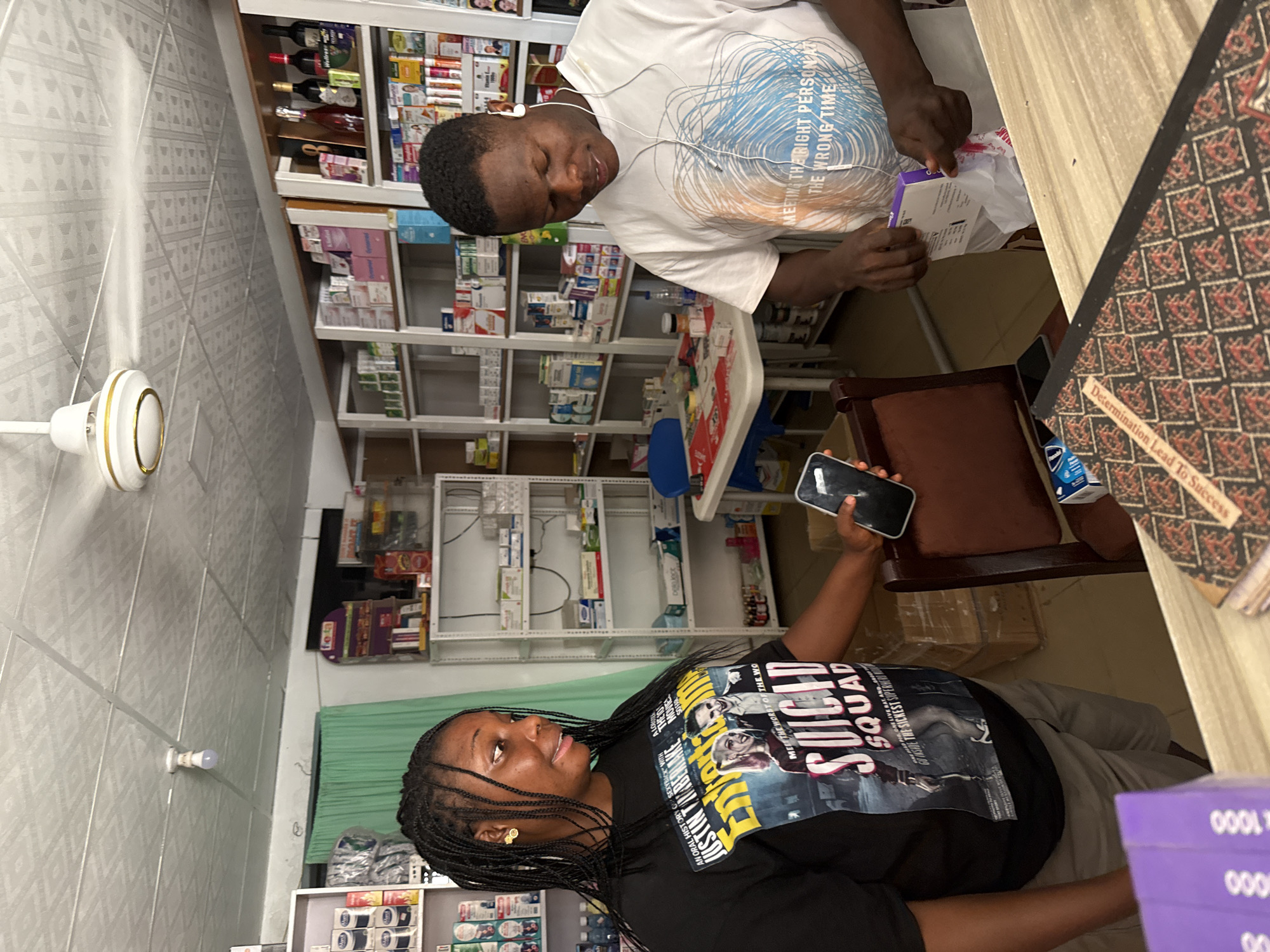 In a pharmacy located in Abuja, Nigeria, a Black woman with braided hair, wearing a black t-shirt with a "Suicide Squad" graphic, stands to the left. She is holding a smartphone in her right hand and looking to her right. To her right, a Black man wearing a white t-shirt with a circular blue and orange design and the text "MEETING THE RIGHT PERSON AT THE WRONG TIME" is standing behind a counter. He is wearing earbuds and is in the process of opening a small purple and white box, holding a white plastic bag from within. The pharmacy shelves in the background are stocked with various medicines and products. A ceiling fan is visible overhead, and the lighting appears to be from overhead fixtures.