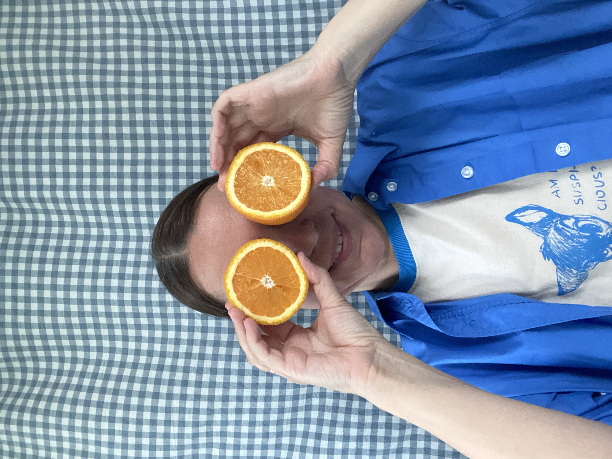 A person lies on a checkered blue and white surface, likely a bedsheet. The person holds two halved oranges in front of their eyes. They are wearing a blue shirt over a white t-shirt with a blue graphic design and the words "AM SUSPICIOUS?". The person's hair is dark and pulled back. The location is likely indoors, but the specific city of Chaikovsky, Russia, cannot be determined from the visual information provided.