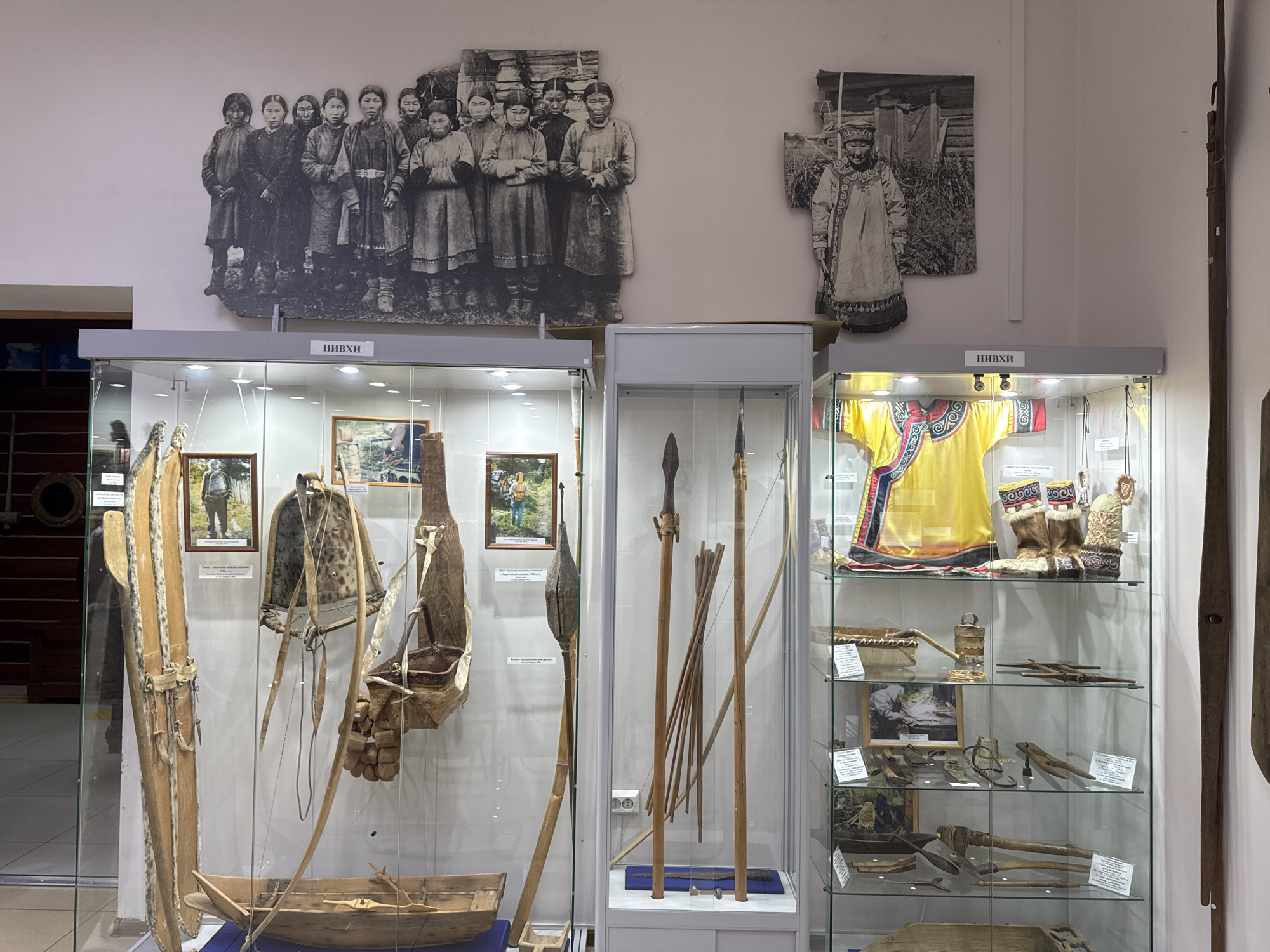 The image depicts an exhibit inside a museum in Poronaysk, Russia, showcasing artifacts and photographs related to indigenous cultures. The display is organized in glass-encased shelves and on the wall.
On the wall, two large black and white photographs are displayed. The one on the left shows a group of women in traditional clothing. The one on the right features a single individual, also in traditional attire, standing outdoors. Below these photographs, two display cases are visible. The left display case contains traditional items such as skis, a bow and arrow, and a boat. The right display case exhibits more cultural artifacts, including clothing, footwear, and tools.
In the foreground, the main focus is on a large, multi-shelf display case. The top shelf holds a brightly colored traditional garment, fur boots, and what appear to be mittens. Below this are several shelves filled with various objects. These include woven baskets, tools, and smaller artifacts, each likely representing a specific aspect of the culture being presented.
The overall environment suggests a museum setting with controlled lighting illuminating the exhibits. The presentation is orderly and aimed at educating visitors about historical and cultural practices.
