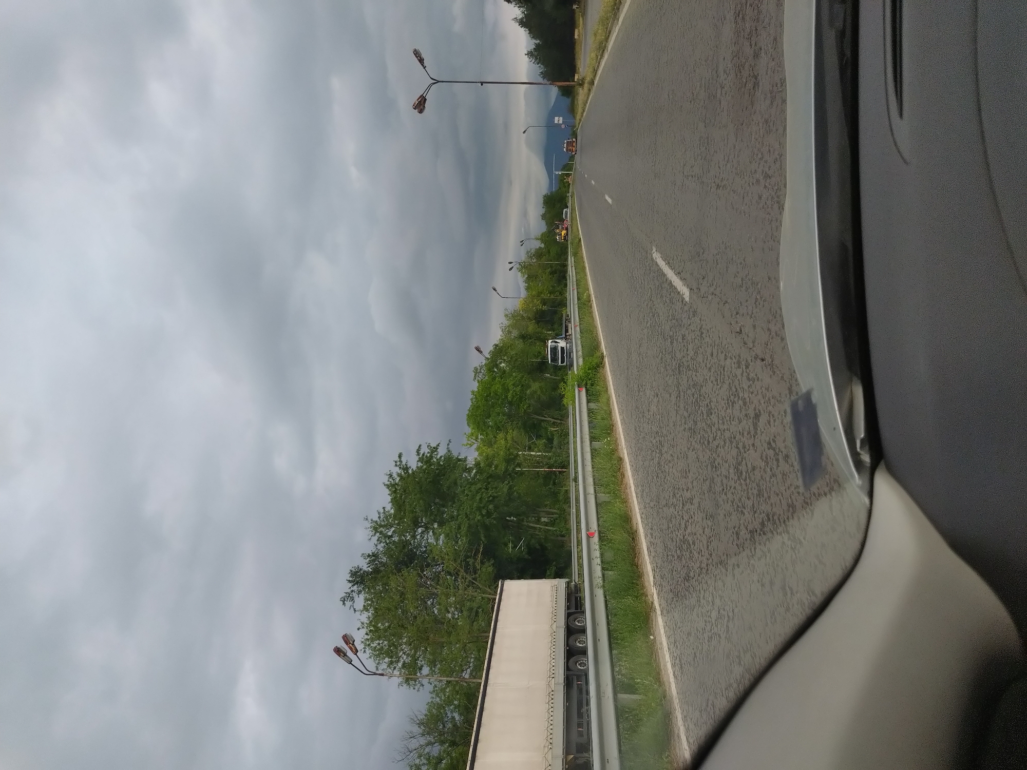 An overcast, cloudy sky looms over a highway in Zmeyovo, Bulgaria. The perspective is from the interior of a vehicle, looking forward through the windshield. A paved road with a white dashed line marking the center stretches into the distance. On the left side of the road, a white semi-trailer truck is parked, and beyond it, a line of lush green trees forms a dense barrier. Several lampposts with multiple lights are visible along the roadside. In the distance, other vehicles, including what appear to be construction vehicles and a truck, are scattered along the highway. The right side of the road is bordered by grass and more trees. The foreground of the image shows the dashboard and part of the windshield frame of the vehicle.
