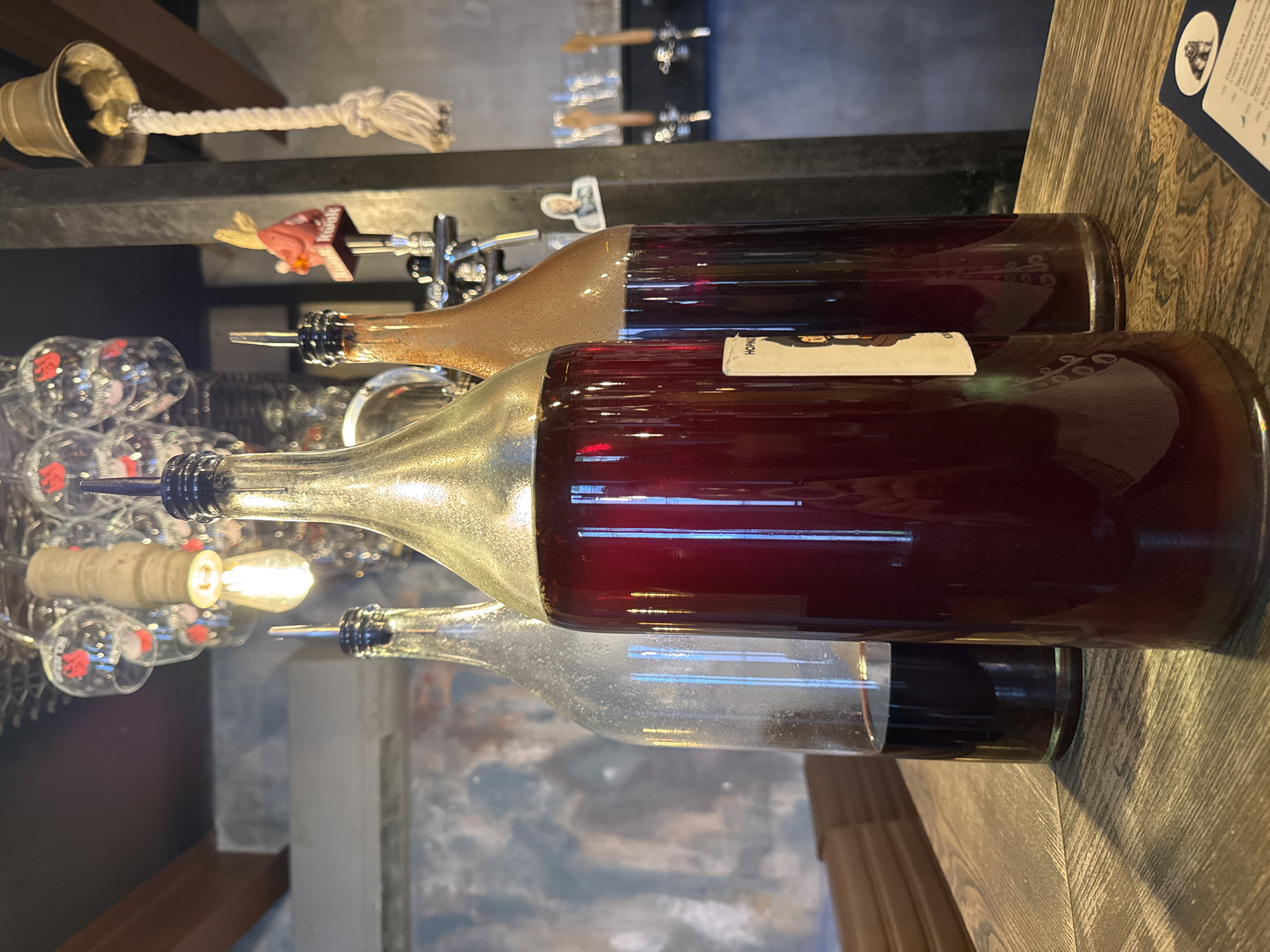 Three large glass bottles containing dark red liquid are displayed on a wooden table. The bottle in the center is the largest and has a metallic gold-colored top with a black pour spout. To its left is a clear glass bottle with a similar black pour spout, containing less liquid at the bottom. To the right of the center bottle is a slightly smaller, amber-colored glass bottle, also with a black pour spout. The background shows a bar or restaurant setting with hanging glasses, a light fixture, a bell, and a beer tap. A label with an illustration is visible on the front of the center bottle. The overall lighting is dim, with a warm tone.