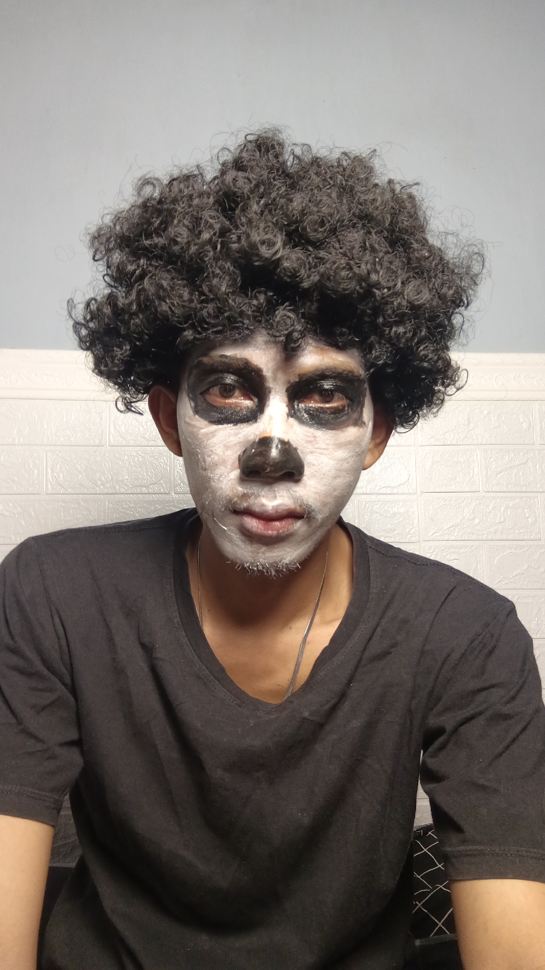 A medium shot captures a person wearing a black t-shirt and a curly black wig, with their face painted white and black. The white paint covers most of their face, with black paint around the eyes and on the nose, resembling a skull or clown makeup. The background consists of a light blue wall above a white textured wall. The image appears to have been taken indoors, possibly in a residential setting in Kebumen, Indonesia, given the information provided. The lighting is consistent with indoor lighting. The subject is looking directly at the camera with a neutral expression.
