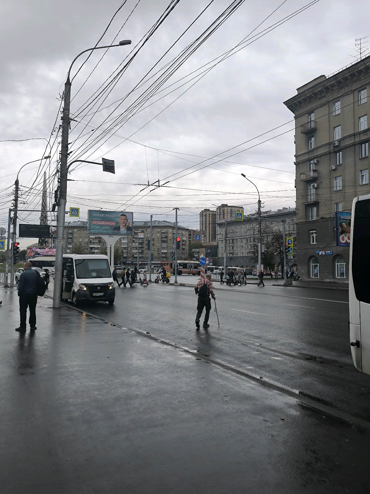 The image depicts a street scene in Novosibirsk, Russia, likely on a cloudy or overcast day given the light and the wet road surface. A white minibus is parked on the right side of the road with its door open, and a man in a dark jacket stands next to it. Further down the road, a person using a walking stick is crossing the street. Pedestrians, some on scooters, are visible at a crosswalk. Numerous overhead wires crisscross the sky, supported by utility poles. Buildings line the street, including a large, multi-story apartment building with balconies and several commercial structures, one of which has a "ВТБ БАНК" sign. Billboards are also present on buildings. The road is wet and reflective, suggesting recent rain. Traffic lights are visible, indicating an intersection.