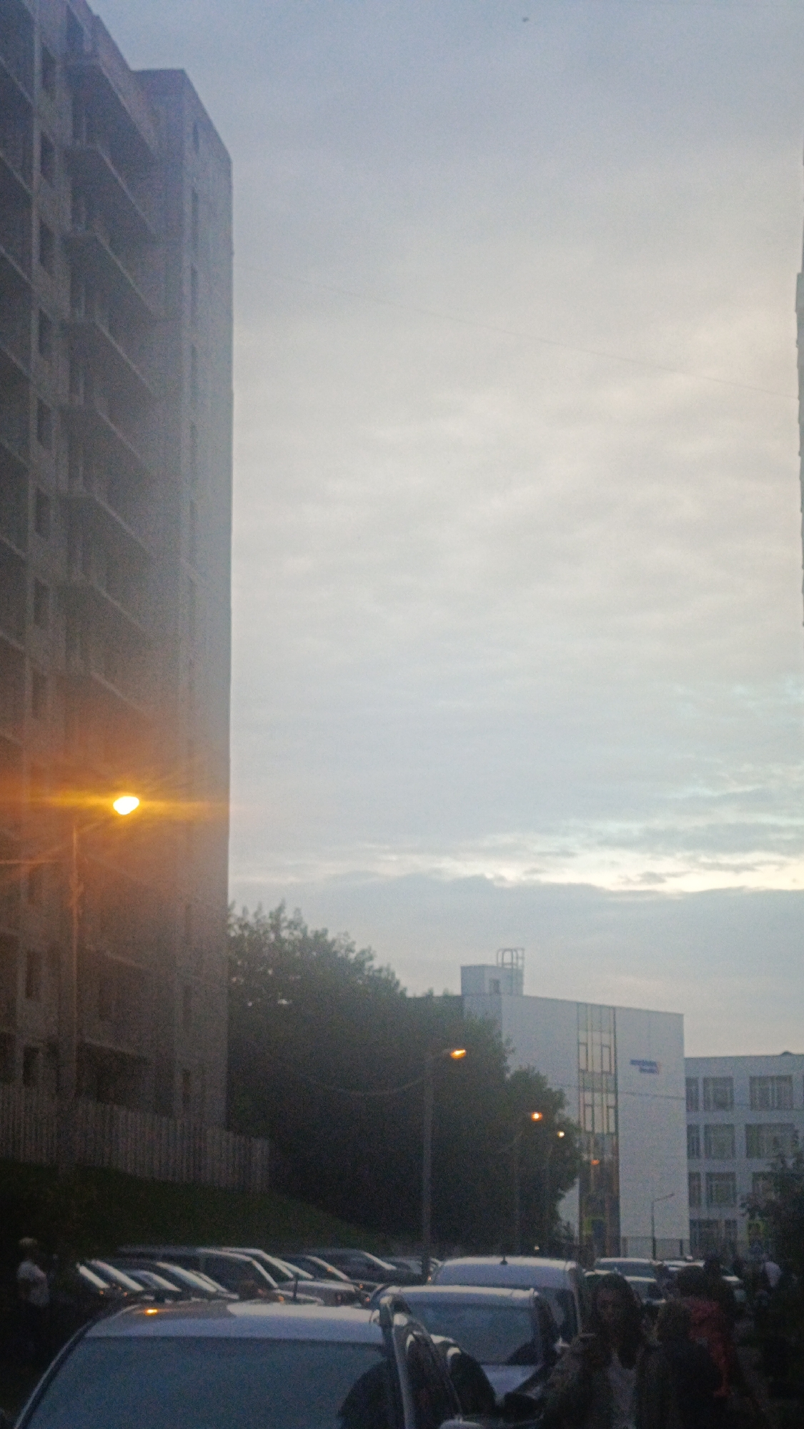 An outdoor, medium shot captures a street scene in Zvenigorod, Russia, under an overcast sky. The left side of the image is dominated by the imposing, unfinished facade of a multi-story concrete building, its raw structure with visible floors and window openings conveying a sense of ongoing construction. A bright orange streetlamp illuminates the lower left portion of this building, casting a warm glow.
To the right of the unfinished building, a line of parked cars is visible, stretching into the distance. Among them are several white vehicles. Further to the right, a modern, white multi-story building with many windows is situated. It appears to be an office or residential complex. Trees are interspersed throughout the scene, particularly in the mid-ground, their foliage dark against the lighter sky.
In the foreground, the tops of several cars are visible, indicating a parking area or street with vehicles. The overall impression is one of an urban environment, likely a residential or mixed-use area, during daylight hours, possibly early morning or late afternoon given the diffused light. The sky is a pale, hazy blue with subtle cloud formations. There are faint indications of people in the lower left and right portions of the image, appearing as silhouettes or indistinct figures, suggesting some pedestrian activity in the vicinity.