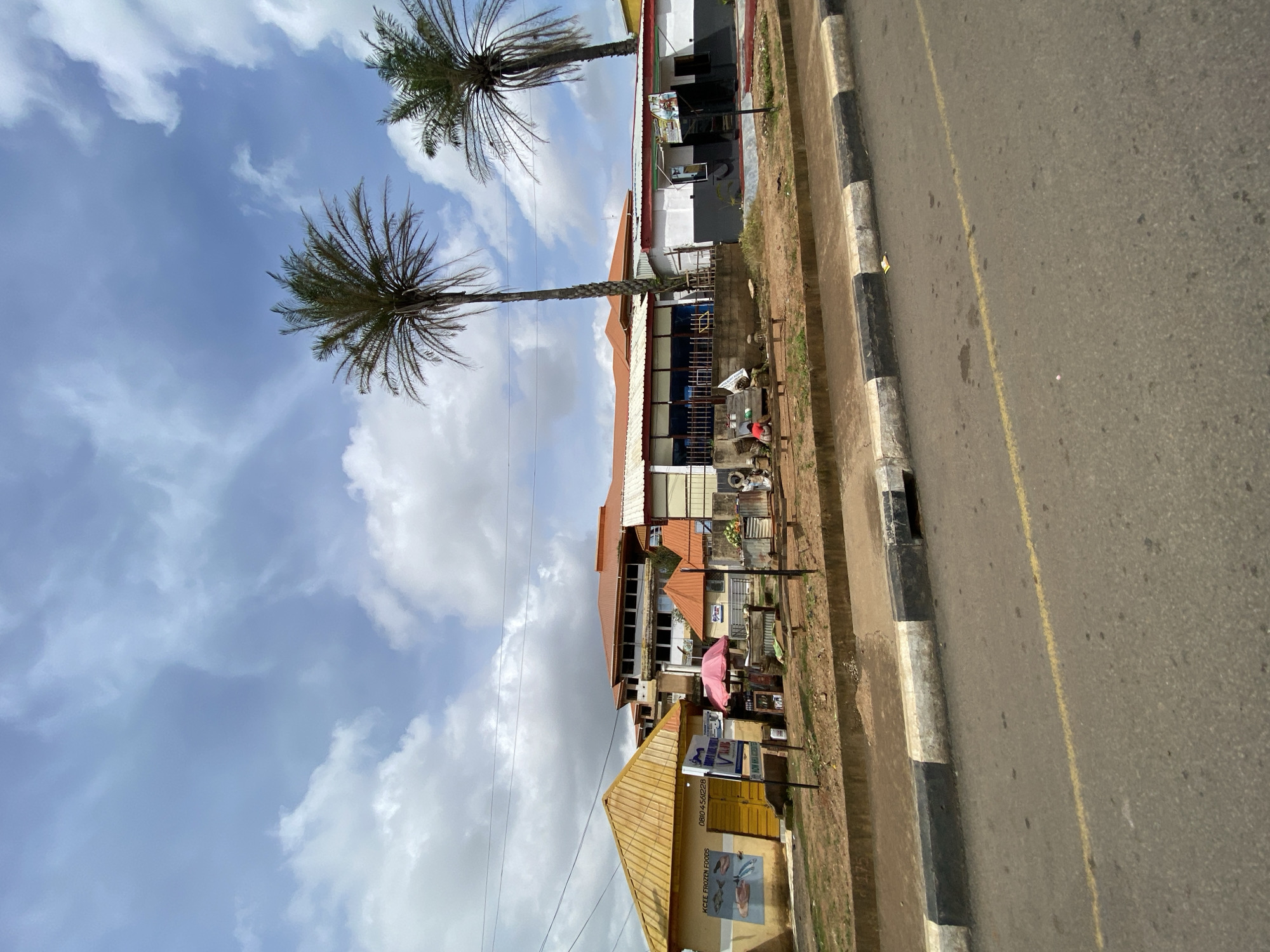 The image shows a street view in Jos, Nigeria. In the foreground, a paved road with a yellow dividing line runs horizontally across the bottom of the frame. A concrete curb, painted with alternating black and white stripes, borders the road. Beyond the curb is a patch of reddish-brown earth, followed by a sidewalk.
The middle ground features a collection of small buildings and structures. To the left, a yellow building with a gabled roof has "KCEE FROZEN FOODS" painted on its side, along with an image of fish. Next to it, a smaller structure with an orange roof has a pink umbrella and various items displayed. A larger, multi-story building with an orange roof dominates the background, with a facade that includes windows and balconies. Two palm trees stand tall in front of this building. Further to the right, a long, single-story building with a white and dark gray exterior is visible, with an entrance and signage.
In front of the buildings, several people are present. Two individuals are seated on a bench, and another person appears to be standing or walking. Other objects visible include benches, crates, and a metal barrier. The sky above is blue with scattered white clouds. The overall scene depicts a commercial area with a mix of shops and a larger building, set against a backdrop of palm trees and a cloudy sky.