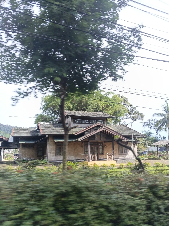 The image captures a rural or semi-rural scene, likely taken from a moving vehicle due to the motion blur in the foreground. The primary subject is a single-story house with a prominent, dark tiled roof and a distinct, elevated front entrance featuring wooden pillars and railings. The house exterior appears to be made of a light brown or beige material, possibly stucco or rough plaster, with dark wooden window frames and doors. A taller section of the house with windows is visible towards the rear.
In the immediate foreground, blurred green bushes and foliage obscure the lower part of the scene, while a dark-barked tree with dense green leaves stands centrally, partially obstructing the view of the house and extending towards the top of the frame. Several black power lines stretch horizontally across the upper portion of the image.
Surrounding the house are various green trees and plants, contributing to a lush environment. To the far right of the house, a simple open-sided structure, possibly a shed or gazebo with a dark roof, is visible. In the distant background on the left, faint outlines of mountains or hills can be seen under a bright, possibly overcast, sky. The time of day appears to be daytime, with ample natural light but no harsh shadows, suggesting clear weather. No specific activity or people are visible, and there is no discernible text in the image. The setting, with its traditional house style and abundant greenery, is consistent with the provided location context of Carcar, Philippines.