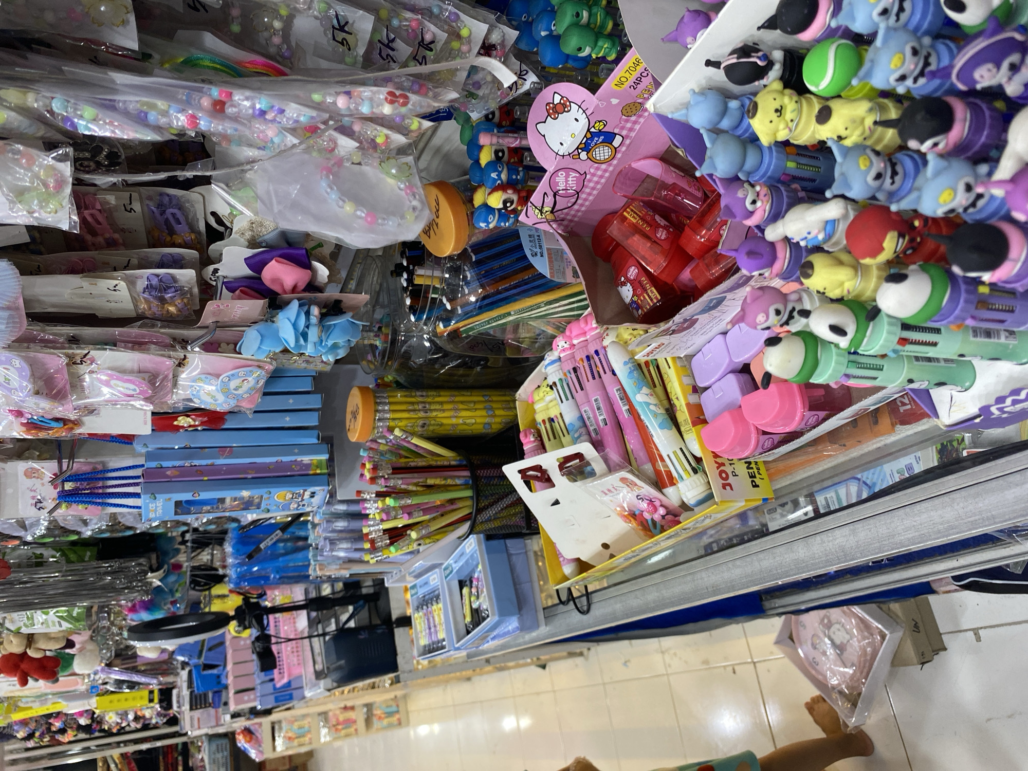 The image shows a stall in a market in Lamteumen Barat, Indonesia, displaying a variety of stationery and small accessories. The stall is densely packed with items arranged on shelves and hanging displays. Prominently featured are colorful pens, pencils, and other writing utensils, many of which have cartoon characters or cute designs. Boxes of colored pencils and multi-colored pens are visible. There are also hair accessories, such as hair clips and headbands, as well as beaded bracelets. Some of the items are packaged in clear plastic, while others are loose. In the foreground, a box displays Hello Kitty themed stationery and collectible figurines of cartoon characters. In the background, more shelves are filled with various products, suggesting a retail environment. The lighting appears to be artificial, typical of an indoor market. A child's leg and foot are visible on the left side of the image, indicating the presence of a customer. The overall impression is one of a vibrant and busy market stall selling a wide assortment of goods.