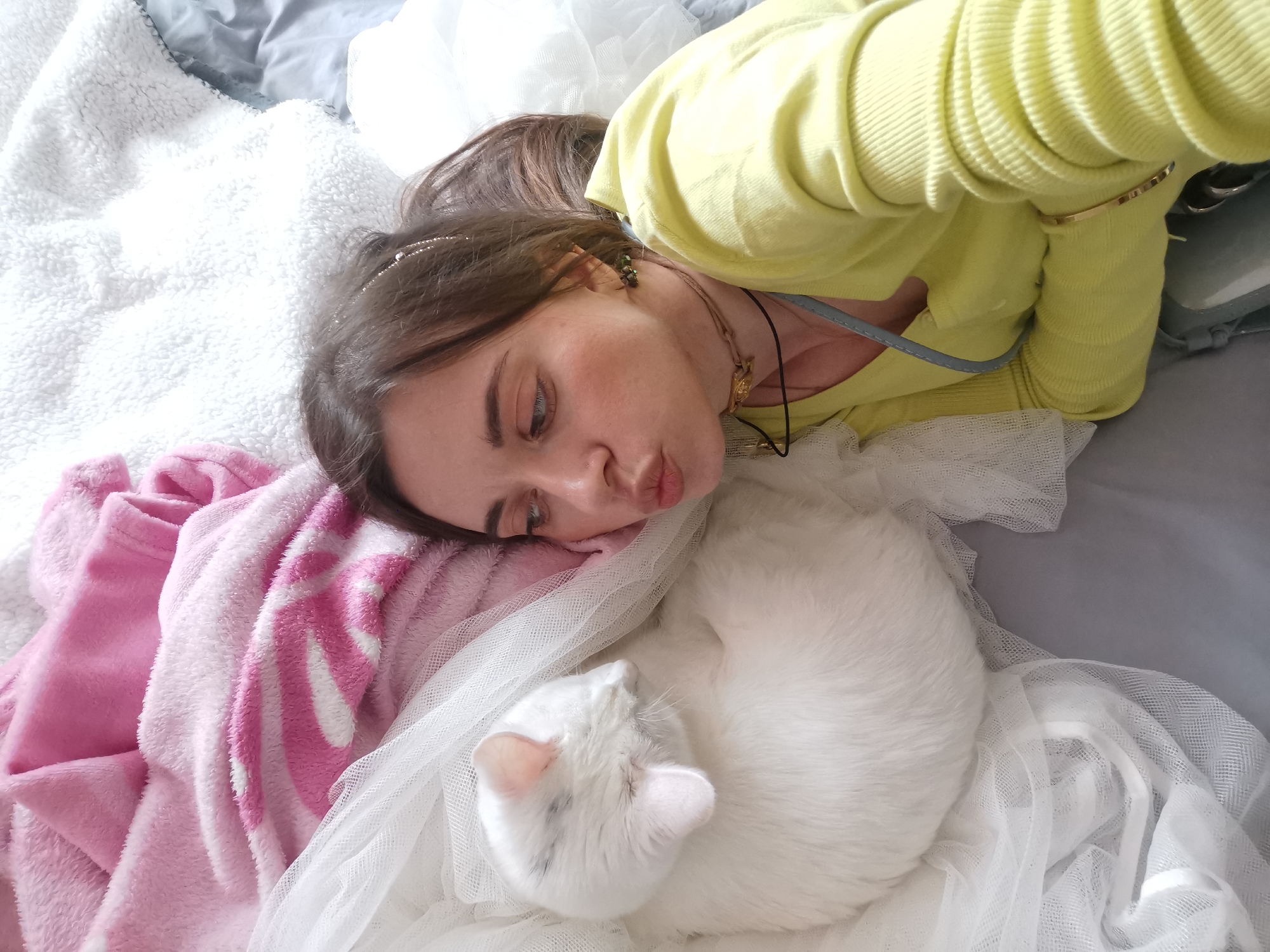 A person is lying on their side next to a white cat curled up on a bed covered in a white tulle fabric. The person has light brown hair and is wearing a yellow sweater. To the left of the person and cat is a pink and white blanket. The background includes a white, textured blanket and a gray sheet. The location is identified as Chaikovsky, Russia.