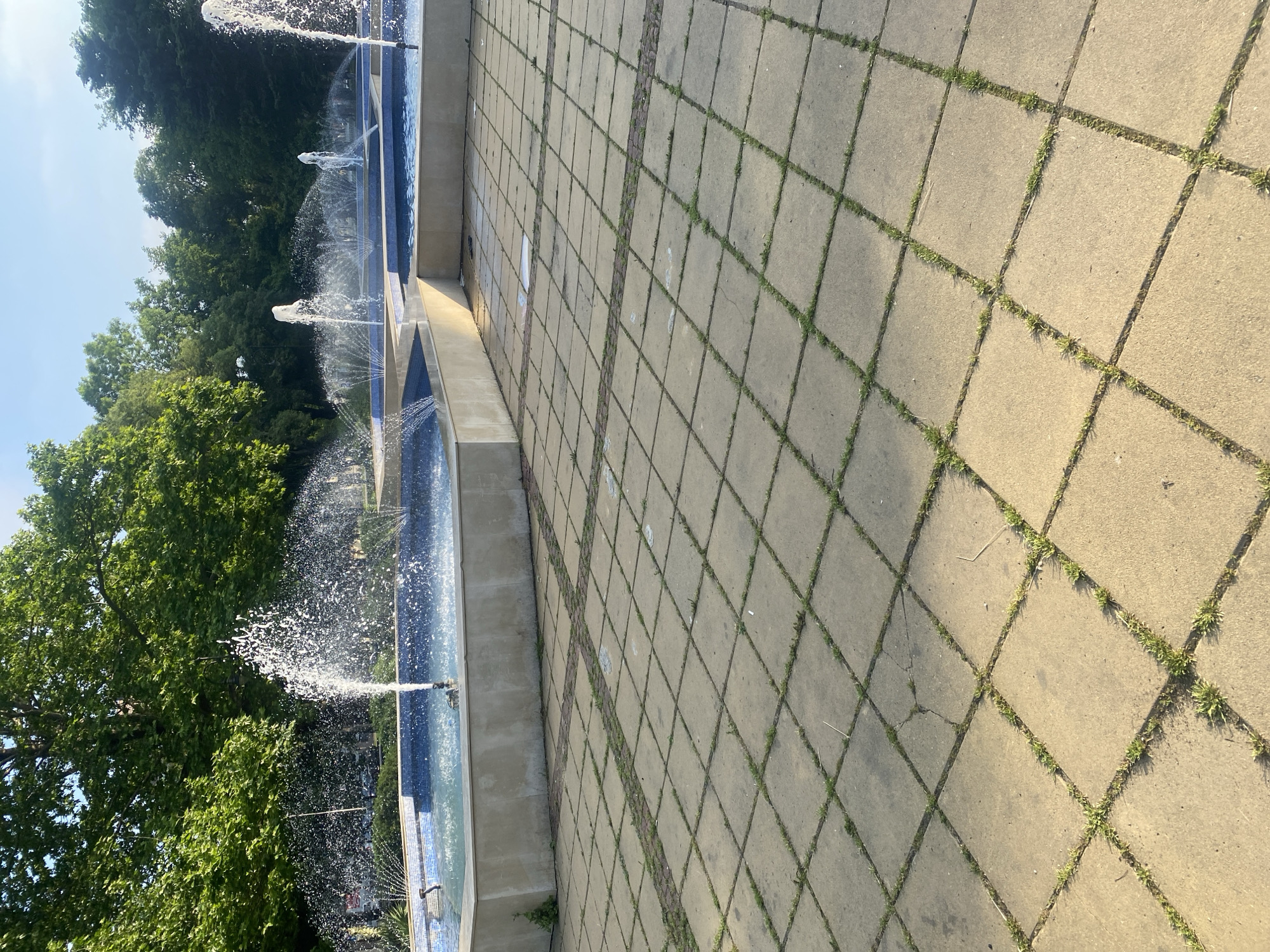 This image depicts the tiered fountain complex located at the entrance to the Sea Garden in Varna, Bulgaria. The foreground features a large paved plaza constructed from light-colored, square paving stones, approximately 50x50 cm, with strips of darker, narrower pavers forming a grid pattern. Abundant green grass and weeds are visible growing in the interstices between the paving stones, indicating a lack of recent maintenance. A significant crack is observable in one of the paving stones in the mid-ground.
Beyond the paved area, a multi-tiered fountain system dominates the middle ground. The fountains are constructed with light beige or off-white concrete walls, and their basins are lined with blue mosaic tiles. Multiple water jets are active, projecting streams of water upwards and outwards, creating visible spray and reflections. Some jets are vertical, while others form a wide, arched spray pattern. Water is visibly circulating within the basins. No individuals are present within the immediate scene.
The background consists of a dense canopy of mature, green deciduous trees, suggesting a park environment. The sky above is clear and light blue, indicating fair weather conditions. Sunlight illuminates the scene, creating distinct shadows on the paved surface and highlighting the water spray. No specific actions or interactions between subjects are occurring. Distant visual elements partially obscured by trees may indicate a road or urban infrastructure beyond the park.