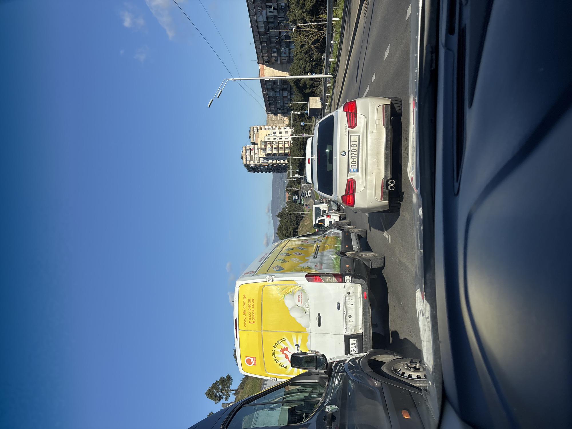 The image shows a congested traffic scene in Tbilisi, Georgia, captured from within a vehicle. A white BMW with the license plate "RO-070-BI" is directly ahead. To the left of the BMW, there's a white van with a yellow advertisement on its side, displaying the web address "www.dila.com.ge" and other Georgian text. The van's license plate appears to end in "3-LA." A dark-colored car is visible in the lower portion of the frame. The background includes residential buildings, trees, a street light, and mountains in the distance under a clear blue sky. The vehicles are stationary or moving slowly in traffic.