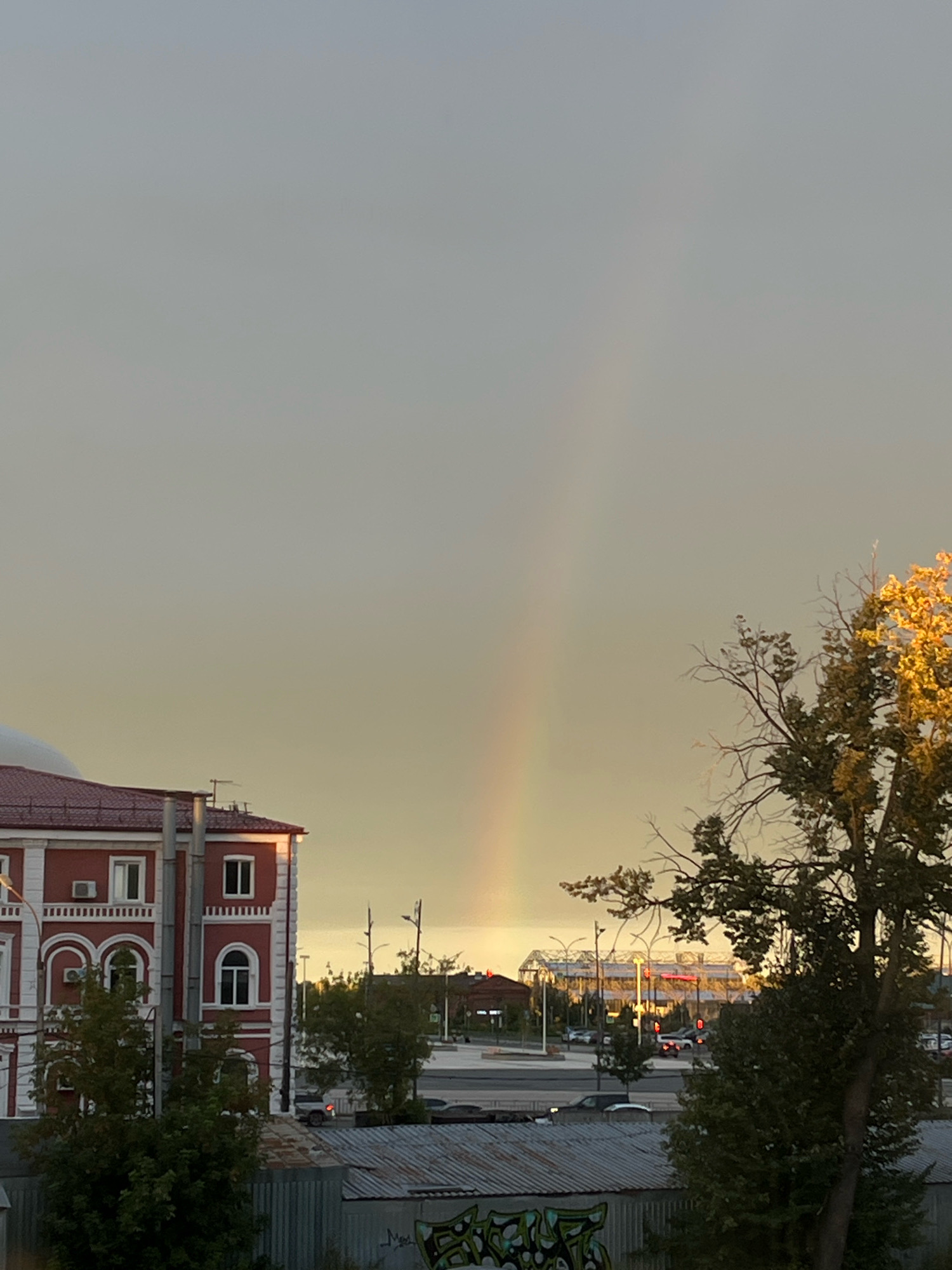 The image shows a faint rainbow in a hazy sky over a city. In the foreground on the left is a red and white building with arched windows. To the right of the building is a tree with yellowing leaves. In the background, there is a modern glass-enclosed structure with illuminated signs and a street with cars. The overall scene suggests an urban environment, possibly in Nizhny Novgorod, Russia, during a time of atmospheric moisture.