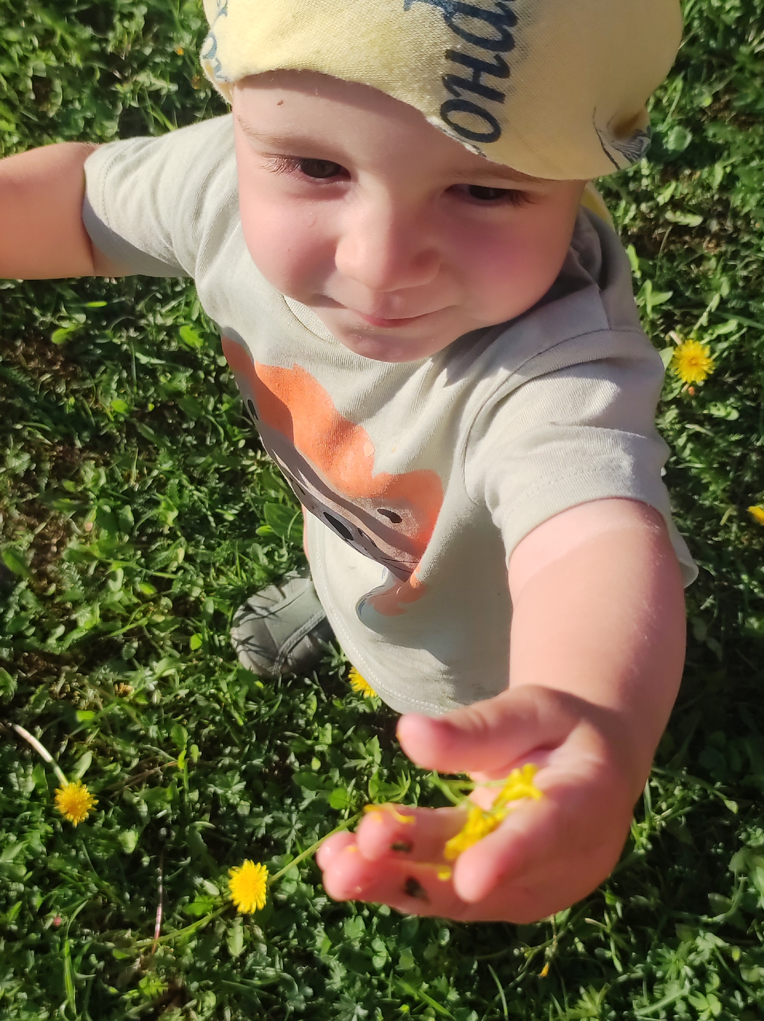 A young child stands in a grassy area, presumably in Фоки, Russia, given the request. The child wears a yellow bandana with Cyrillic writing on their head and a light-colored shirt with an orange design. In their outstretched hand, they hold yellow flower petals. The surrounding environment features green grass dotted with yellow dandelions. A single gray shoe is visible in the background.