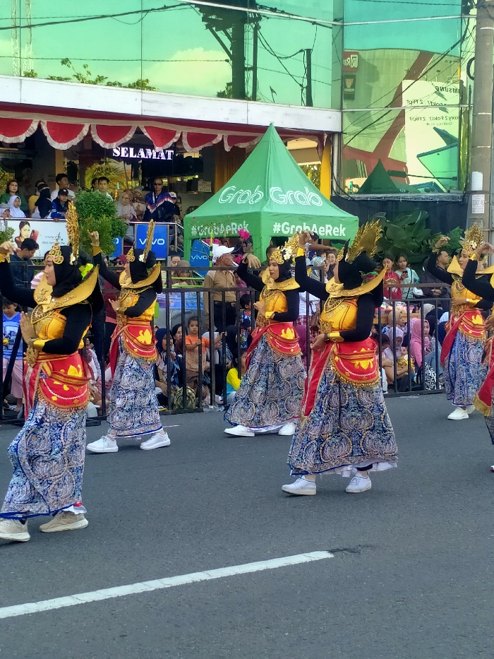 The image displays a public street scene in Madiun, Indonesia, during what appears to be a parade or cultural performance. In the foreground and midground, a line of five female individuals, identified as dancers, are in motion, positioned on an asphalt road featuring a white dividing line. Each dancer wears a costume consisting of a black long-sleeved top, a bright yellow/gold sleeveless bodice, a red and gold patterned hip sash, a long blue skirt with intricate white and gold patterns, an ornate gold headdress, and white athletic sneakers. Their arms are raised in stylized dance poses.
Behind the dancers, a crowd of spectators, varying in age, is gathered behind black metal barriers, observing the event. Some spectators are visible on a raised platform or balcony to the left. In the background, two buildings are prominent: one features a green reflective glass facade, while the other has a white facade with red and white scalloped trim and an illuminated LED sign displaying "SELAMAT." A green 'Grab' branded pop-up tent, also featuring the text '#GrabAeRek', is situated on the right side of the street. The scene is illuminated by natural daylight.
