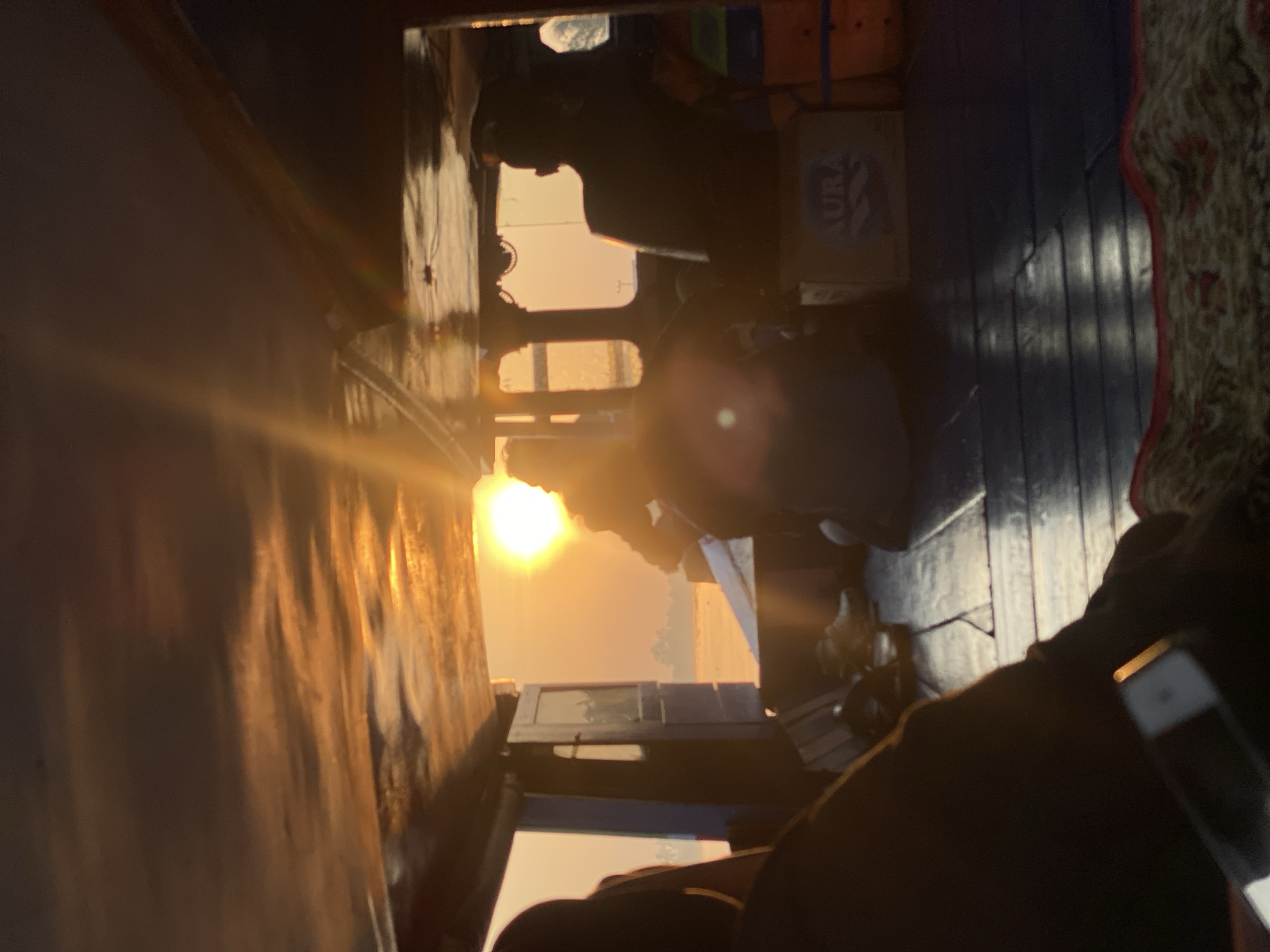 This is an indoor, low-angle, medium shot of a boat interior at sunset, with the sun's glare dominating the right side of the frame. The boat is on the river in Sungai Tandipah, Indonesia. Several individuals are visible, mostly silhouetted against the bright orange sky and sunlight. One person, wearing a hat, is seated in the driver's position, facing forward. Another person is seated to their right. In the foreground, a person's arm is visible, holding a smartphone. The boat has a wooden floor and a dark, sloped ceiling with visible support beams. A cardboard box with the label "AURA" is visible on the right side of the boat. The overall lighting is warm and atmospheric due to the setting sun.