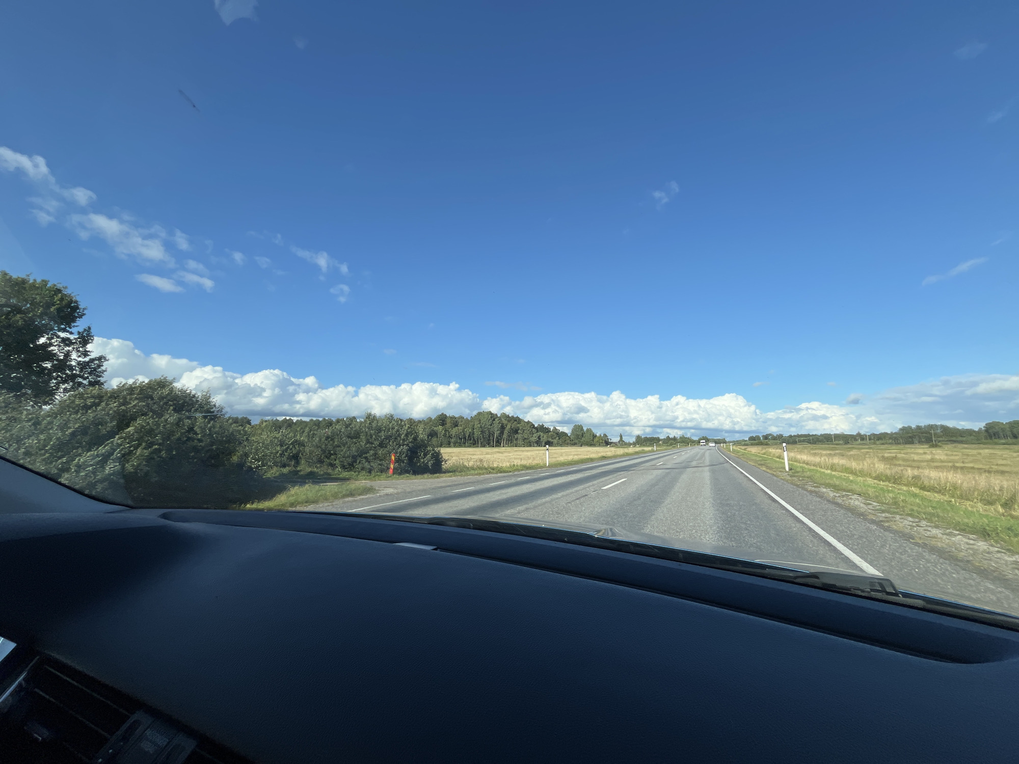 The image captures a view from the interior of a vehicle traveling on a road in Lihula, Estonia. The perspective is from the driver's seat, showing the dashboard in the foreground. The road ahead is paved, with white lane markings visible. To the left of the road, there are trees and vegetation, followed by a field of tall, dry grass. To the right, there is a similar grassy field and a line of trees in the distance. The sky is a clear, vibrant blue with scattered white cumulus clouds. The sun appears to be shining, casting shadows on the road. There are no people visible in the image, but there are a few vehicles in the distance on the road. The overall scene depicts a rural highway landscape under a sunny sky.