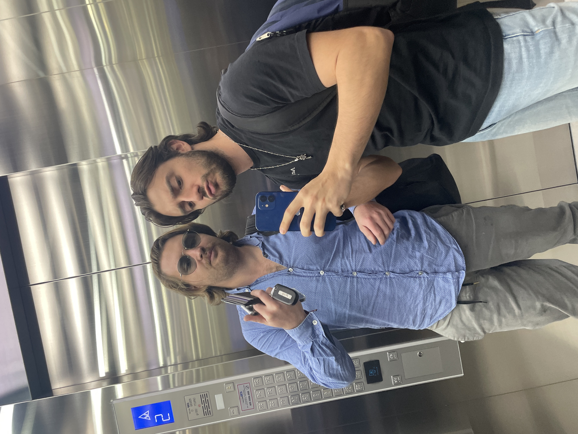 Two men take a selfie in an elevator. One man wears a black t-shirt, light blue jeans, and a silver necklace. The second man wears sunglasses and a light blue button-down shirt, gray slacks, and holds keys in his hand. The elevator panel indicates it is in Huixquilucan de Degollado, Mexico. The elevator walls appear to be brushed metal.