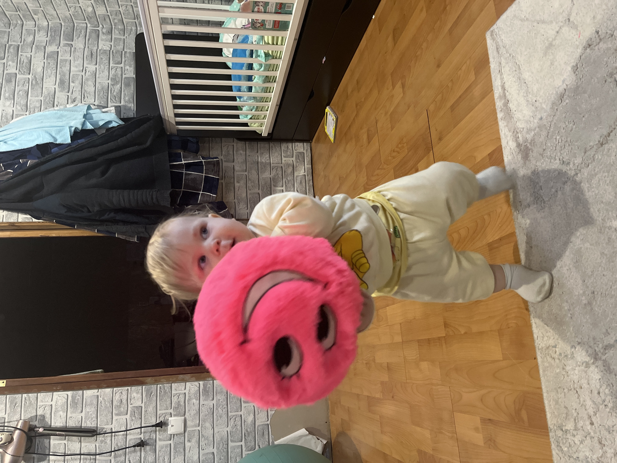 A vertically oriented indoor photograph captures a young child holding a large, bright pink, fluffy smiley face plush toy. The child, with light-colored hair and fair skin, is positioned centrally, looking directly at the camera with a neutral expression. They are wearing a light-colored (possibly cream or yellow) long-sleeved onesie or pajamas featuring a yellow cartoon character print, along with white socks.
The scene depicts a residential room in what is understood to be Vesyoloye, Russia. The background features a wall adorned with grey brick-patterned wallpaper or tiles. To the left, a white crib with visible railings is partially present, alongside a dark piece of furniture and a stack of clothing. The floor is covered partly by light wooden planks, indicative of laminate or parquet flooring, and partly by a light-colored, textured rug on the right. In the bottom left corner, a portion of a green exercise ball is visible, along with a white electrical outlet on the brick wall, where a power cord is plugged in. A dark screen or surface is also partially visible near the outlet. On the wooden floor, a small yellow item, possibly a toy or book, can be seen. The lighting suggests an indoor setting, likely with artificial light, without specific cues for the time of day. Text on a package inside the crib is present but illegible due to blurriness.