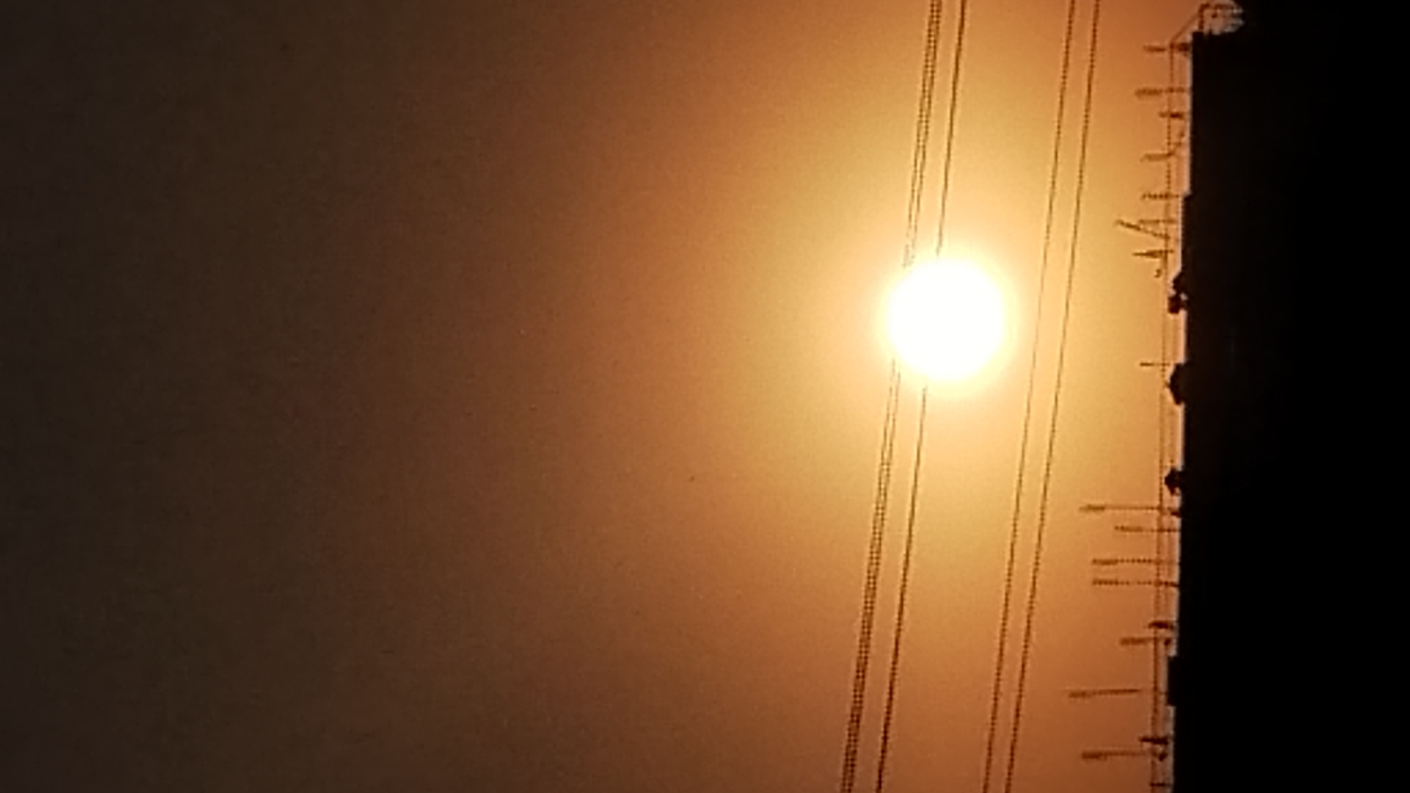 This is a photograph of a sunset in the city of Unknown, Unknown. The sun is a bright, glowing orb in the sky, surrounded by a haze of orange and yellow. Several power lines stretch across the frame, silhouetted against the vibrant sky. Below the power lines, the dark silhouettes of buildings and structures are visible, suggesting an urban landscape. The overall scene evokes a sense of twilight and the end of the day.