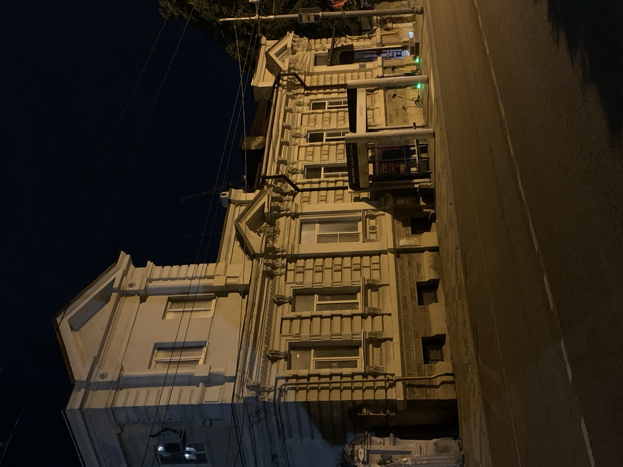 The image shows a multistory building illuminated by streetlights at night. The building has an ornate facade with several windows and decorative elements. To the right of the building is a dark street. Overhead electrical lines are visible against the dark sky. The location in the city is unknown.