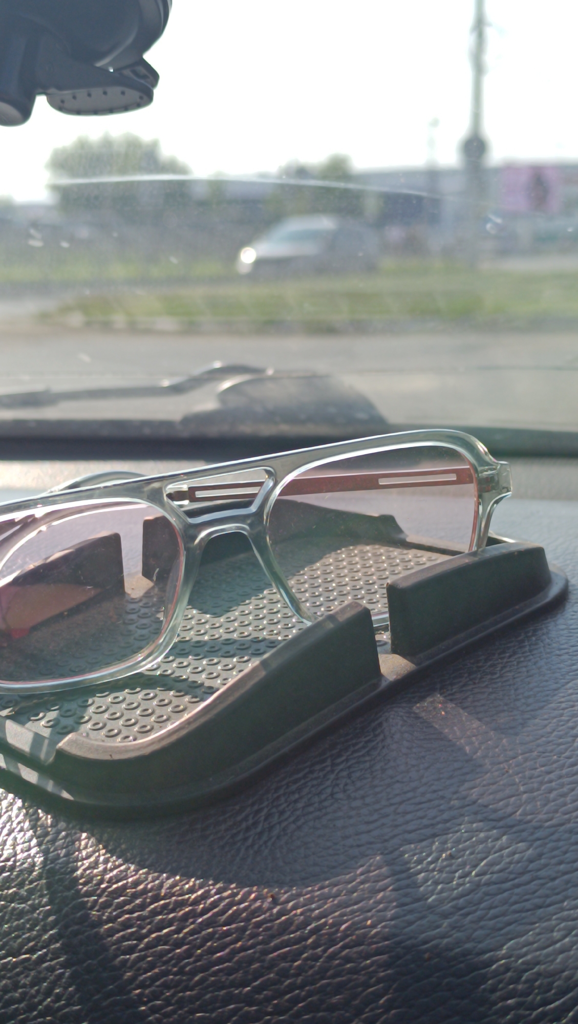 The image shows a pair of clear-rimmed sunglasses with pinkish lenses resting on a black, textured dashboard inside a vehicle. The sunglasses are positioned on a black, non-slip mat designed to hold objects. The dashboard is made of a dark, textured material that appears to be leather or a similar synthetic. Sunlight is casting shadows on the dashboard, indicating it is daytime. In the background, seen through the windshield, is a blurred view of an outdoor scene, possibly a road with trees and a building. There is no clear indication of the specific location within Chaikovsky, Russia, beyond the context provided of being inside a vehicle.