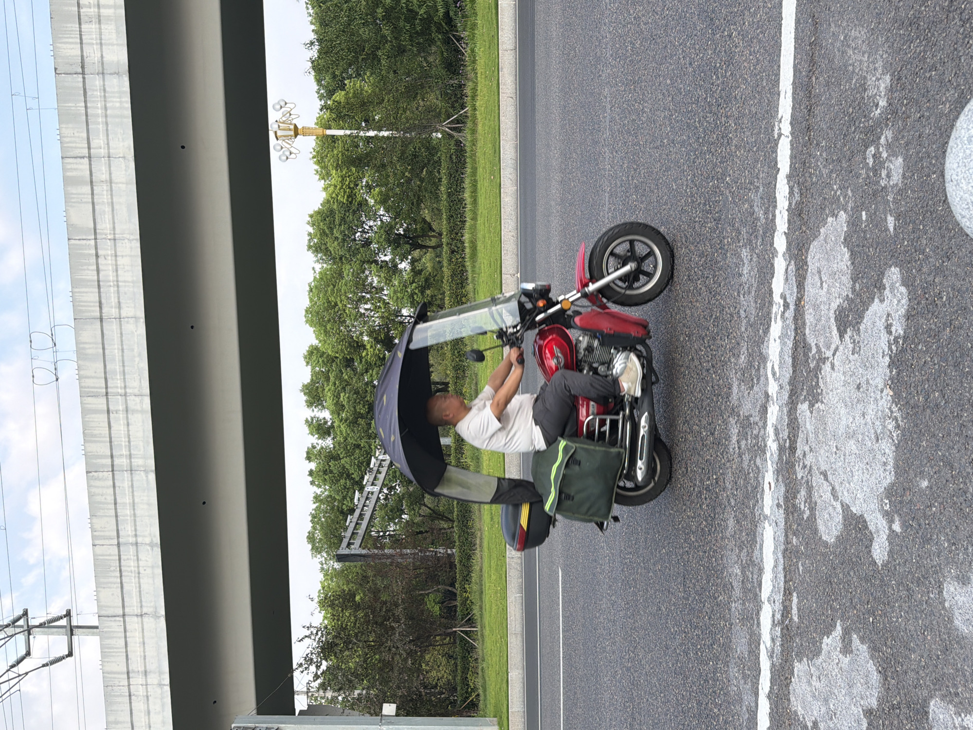 A man is riding a red motorcycle with a black umbrella for shade and a transparent windscreen on a paved road. He is wearing a white t-shirt and dark pants, with beige shoes. The motorcycle has a black storage box on the back and two green saddlebags. In the background, there is a concrete overpass with power lines, a line of trees, and a lamppost. The road has a white line marking. The image appears to have been taken in the city of Jiangjin, China.