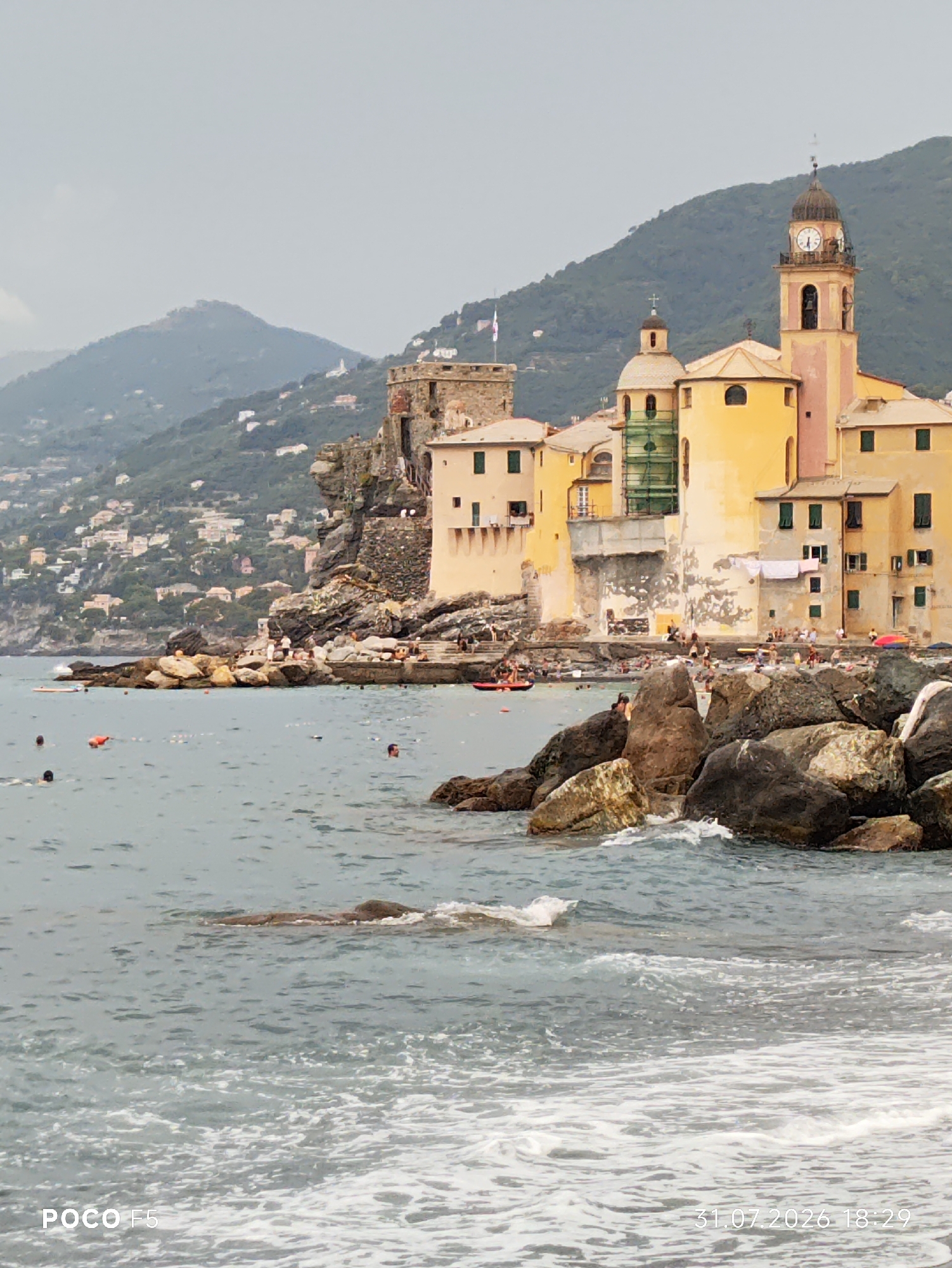 This image depicts a coastal scene in Camogli, Italy, showcasing the iconic Basilica di Santa Maria Assunta. The basilica, a prominent structure with a tall clock tower and pastel yellow and pink facade, stands on a rocky outcrop overlooking the Ligurian Sea. Multiple smaller buildings, also painted in warm, sun-bleached tones, are clustered around it, characteristic of the colorful architecture found in Italian coastal towns.
In the foreground, the sea is choppy with waves crashing against a breakwater made of large, dark rocks. Several people are visible in the water, some swimming and others wading near the shore. Others are gathered on the stone embankment and stairs leading up to the buildings. A small red inflatable raft is present in the water, and further out, buoys are scattered across the sea. The background is dominated by a lush, green hillside dotted with more houses, suggesting a densely populated coastal community extending up the slopes. The sky is overcast, casting a soft, diffused light over the scene. A digital timestamp in the lower right corner indicates the image was taken on "31.07.2026 18:29".