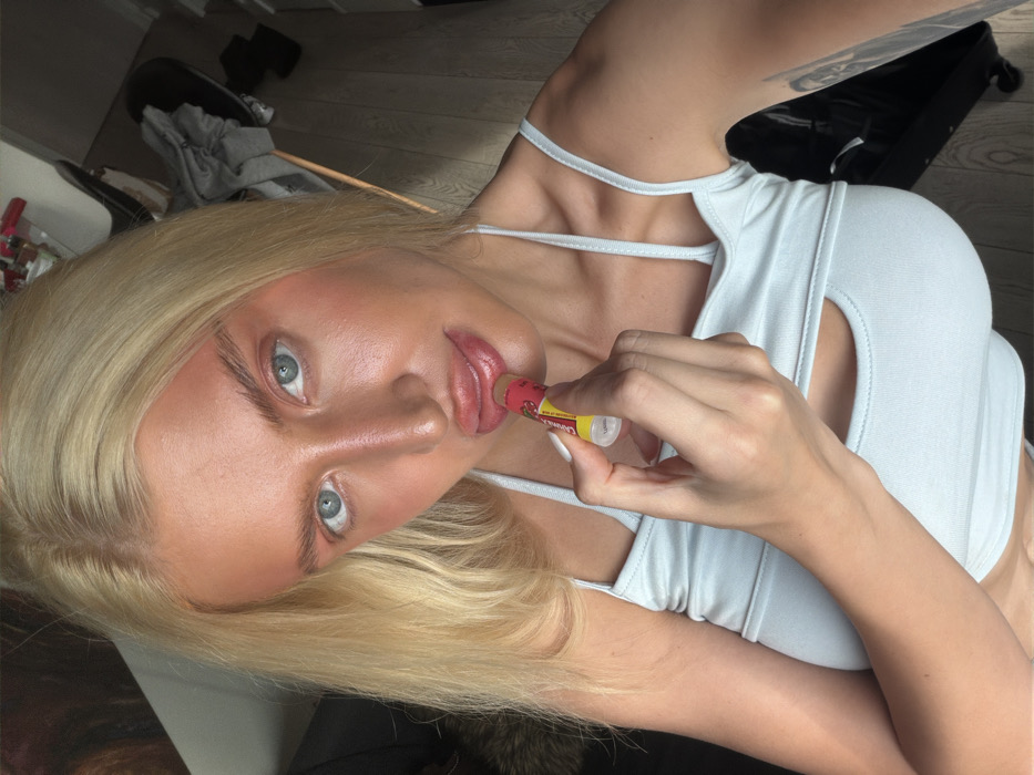 A fair-skinned woman with long blonde hair and blue eyes is depicted in a close-up, chest-up shot. Her face is made up with a noticeable orange-red blush or bronzer across her forehead, cheekbones, and nose. Her lips are full and glossy. She is actively applying a white-tubed lip balm, labeled "Cranberry," to her upper lip with her right hand, holding the balm with her thumb and forefinger. She wears a light blue or white sleeveless top featuring thin straps and a horizontal cut-out below the bust line. A dark tattoo is partially visible on her left upper arm. The background is an indoor environment, showing a light-colored wooden floor, a black office chair draped with grey clothing, and indistinct objects further in the distance. The specific location within the city of Belgrade, Serbia, cannot be ascertained from the image's visual context.
