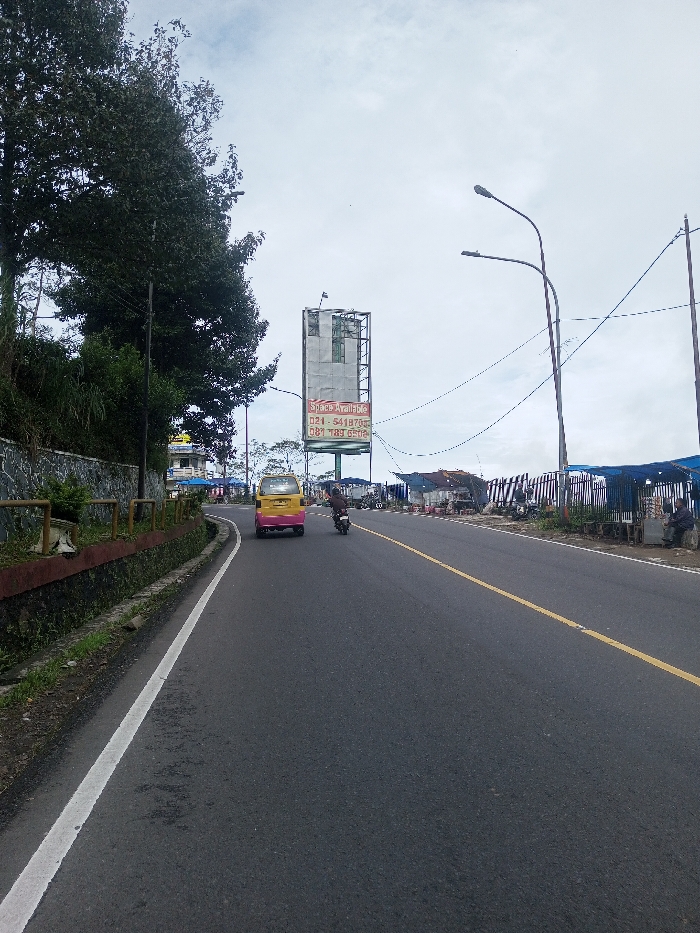 The image presents an overcast street scene in Sandamal Eliya, Sri Lanka, viewed from a low angle along the direction of travel.
In the foreground and extending along the left, a dark asphalt road features a solid white line delineating its edge, alongside a narrow concrete drainage ditch and a low concrete wall topped with yellow metal railings. Behind this wall, dense green foliage and large trees line the roadside. Partially visible further back on the left is a light-colored building with a prominent domed roof.
On the road, a yellow and pink minivan-like vehicle is traveling away from the viewer in the left lane. A single individual on a motorcycle, wearing dark clothing, follows in the right lane, also moving away from the camera.
The right side of the road is bordered by a series of informal market stalls covered by blue tarpaulins. Various goods are displayed on tables or stands in front of these stalls. Two individuals are seated on low wooden stools at the far right edge, near one of the stalls. A tall metal billboard structure with a sign reading "Space Available" and displaying Indonesian contact numbers ("021-541870X," "081-789 650X") stands prominently on the right. Two grey street light poles with double luminaires are also visible on the right, with overhead utility wires crossing the scene. The sky is uniformly overcast.