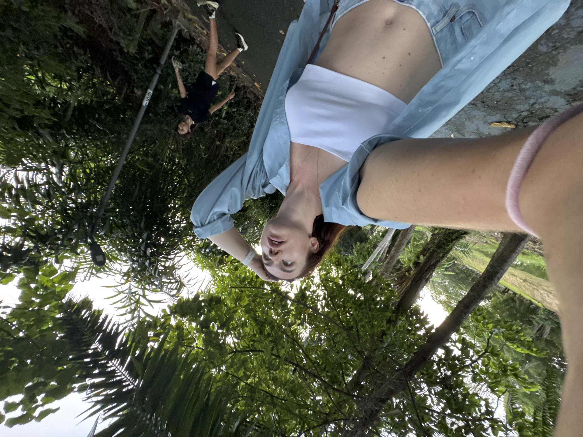 The image depicts two individuals outdoors, likely in a park or a natural setting within Phuket City Municipality, Thailand. The foreground is dominated by a woman taking a selfie. She has auburn hair and is looking upwards with her right hand resting on her head. She is wearing a light blue open shirt over a white crop top and denim shorts. Her left arm is visible, adorned with a pink hair tie.
In the background, a second person, appearing to be a man, is captured in motion, possibly jogging or exercising on a paved path. He is wearing a black t-shirt and black shorts, with athletic shoes. The environment is lush with dense green foliage and trees framing the scene. A street lamp is visible above and to the left of the man. The lighting suggests it is daytime, with soft natural light filtering through the trees. The overall impression is of an active, outdoor moment amidst a verdant urban landscape.