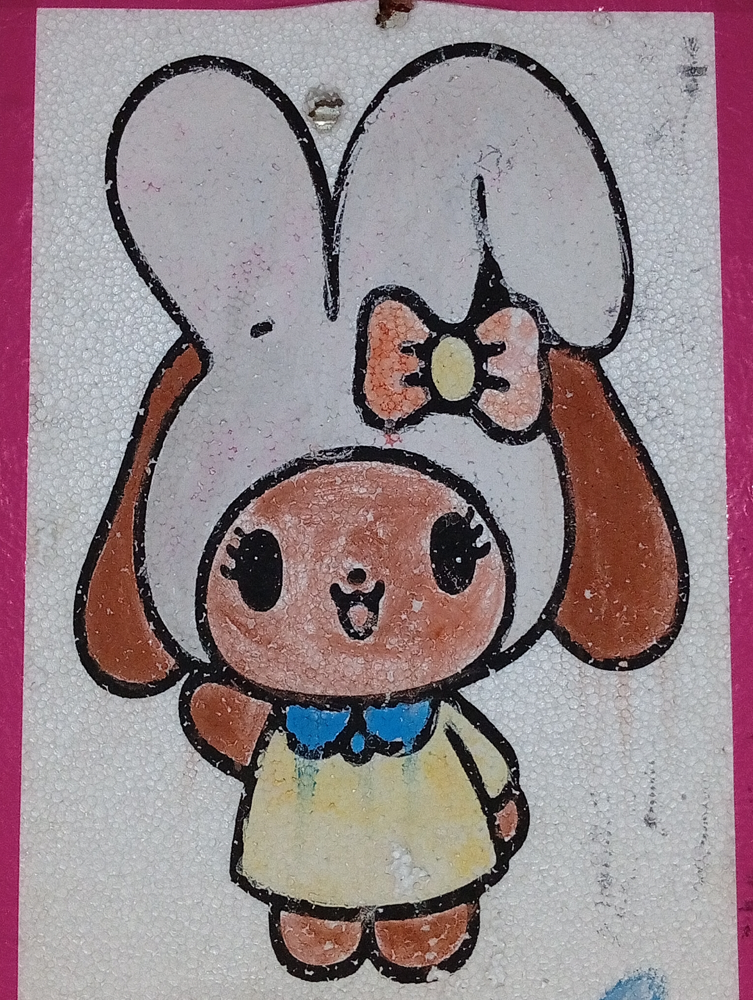 The image shows a drawing of a cartoon bunny character on a white, textured surface, possibly styrofoam. The bunny is wearing a large hat that resembles bunny ears, a bow on top, and a yellow dress with a blue collar. The character has large black eyes, a small mouth, and brown accents on its ears and feet. The drawing is outlined in black. The background is white and appears to be a material with a granular texture. The edges of the drawing are bordered by a bright pink material. There is no visible indication of the city of Lhokseumawe, Indonesia, in this image.