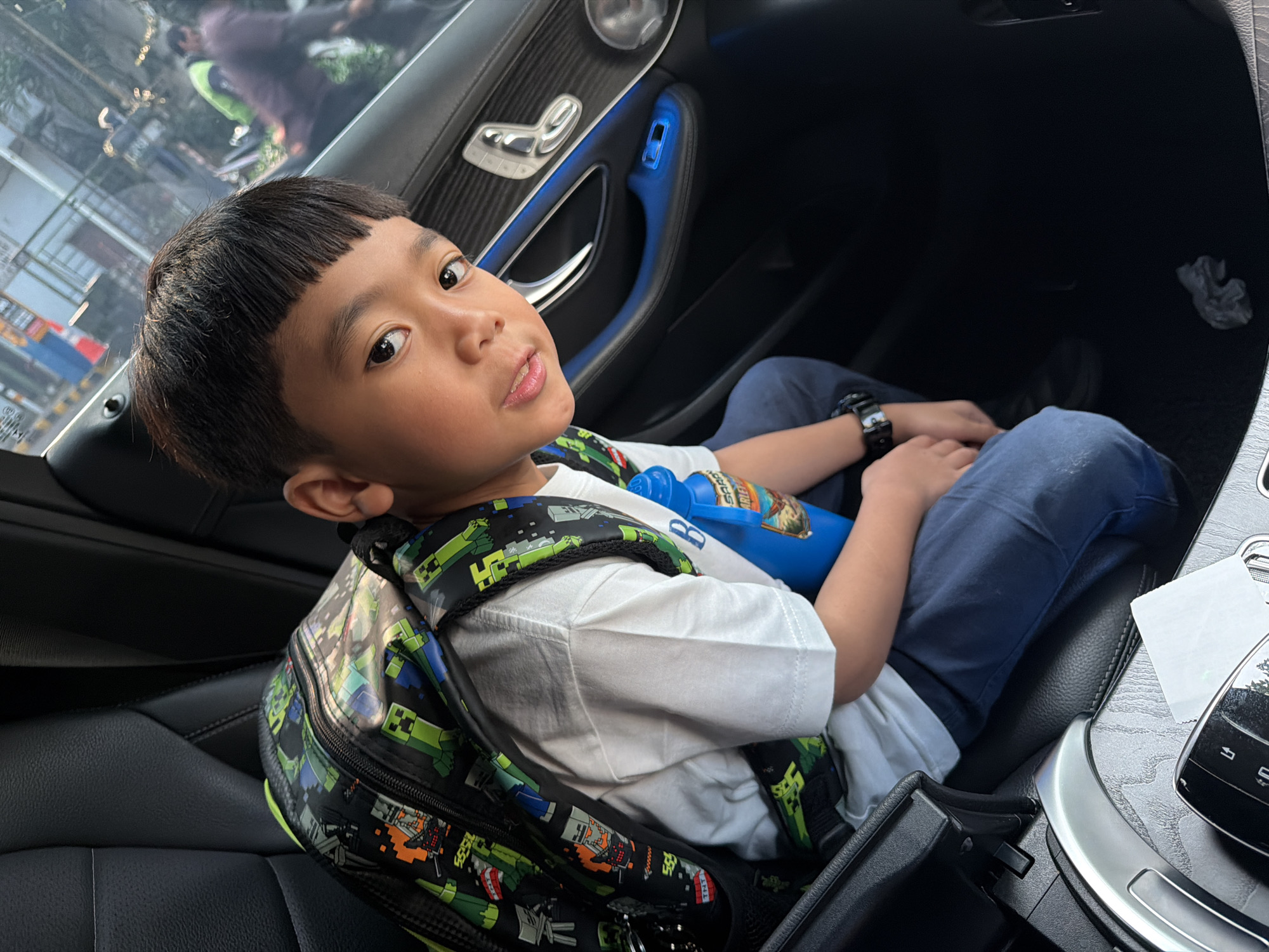 In Central Jakarta, Indonesia, a young boy with short, dark hair and bangs is seated in the back of a car. He is wearing a white t-shirt and dark blue pants, with a black backpack featuring a Minecraft-themed design adorned with green pixelated characters and orange TNT blocks. A blue water bottle with a label depicting a bird and the name "Sarah" is positioned next to him. The car's interior is visible, with black leather seats and accents of blue ambient lighting around the door panel. The boy is looking directly at the camera, his expression neutral. Outside the car, reflections in the window show a blurred street scene with vehicles and pedestrians, suggesting an urban environment.